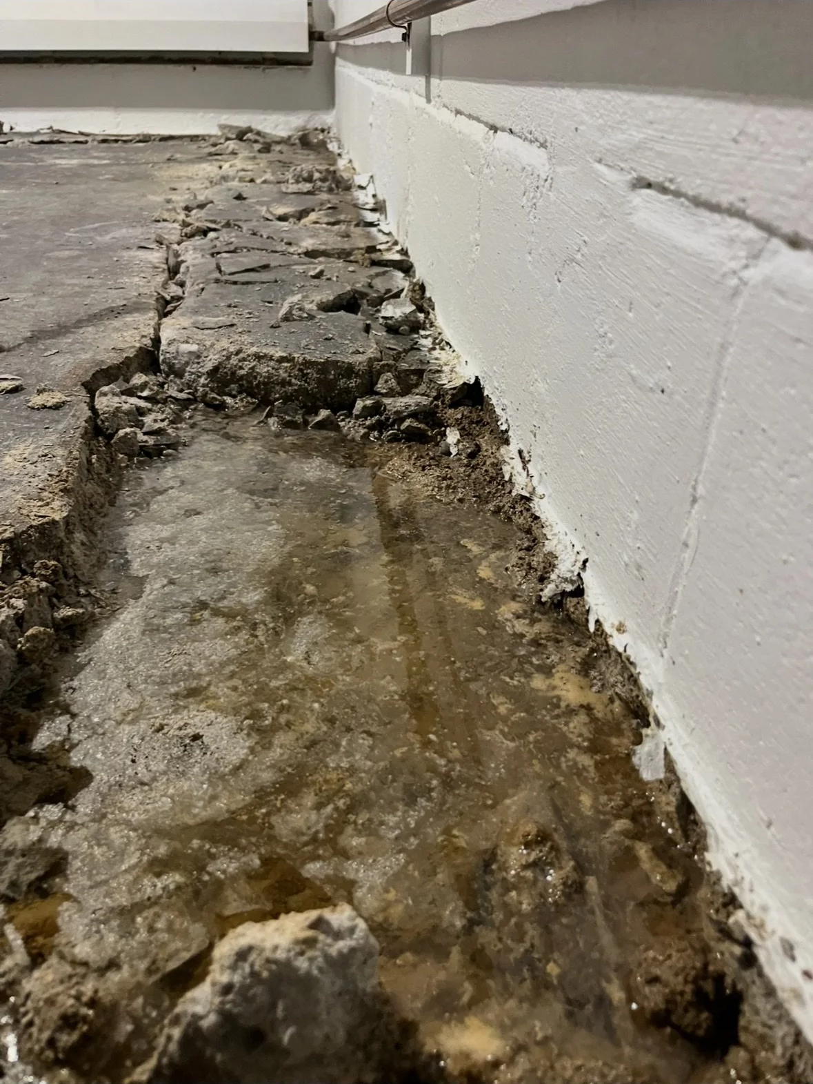 Close-up of a construction site showing a concrete floor with a trench filled with water, surrounded by rocks and dirt, next to a painted white brick wall.