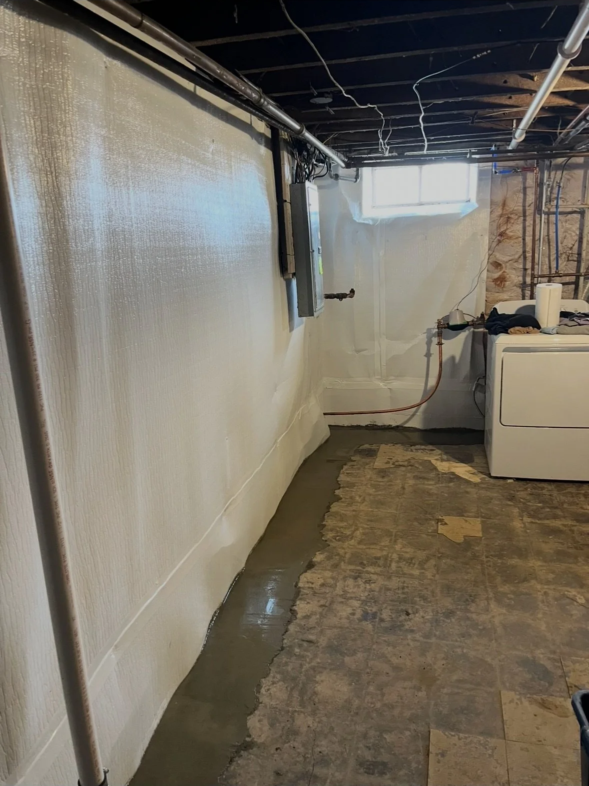 A basement laundry area with unfinished ceiling and a window. The wall is partially painted or coated, with a level of wet concrete or patch along the bottom. A white washing machine is placed against the right wall, with laundry clothes on top. Pipi