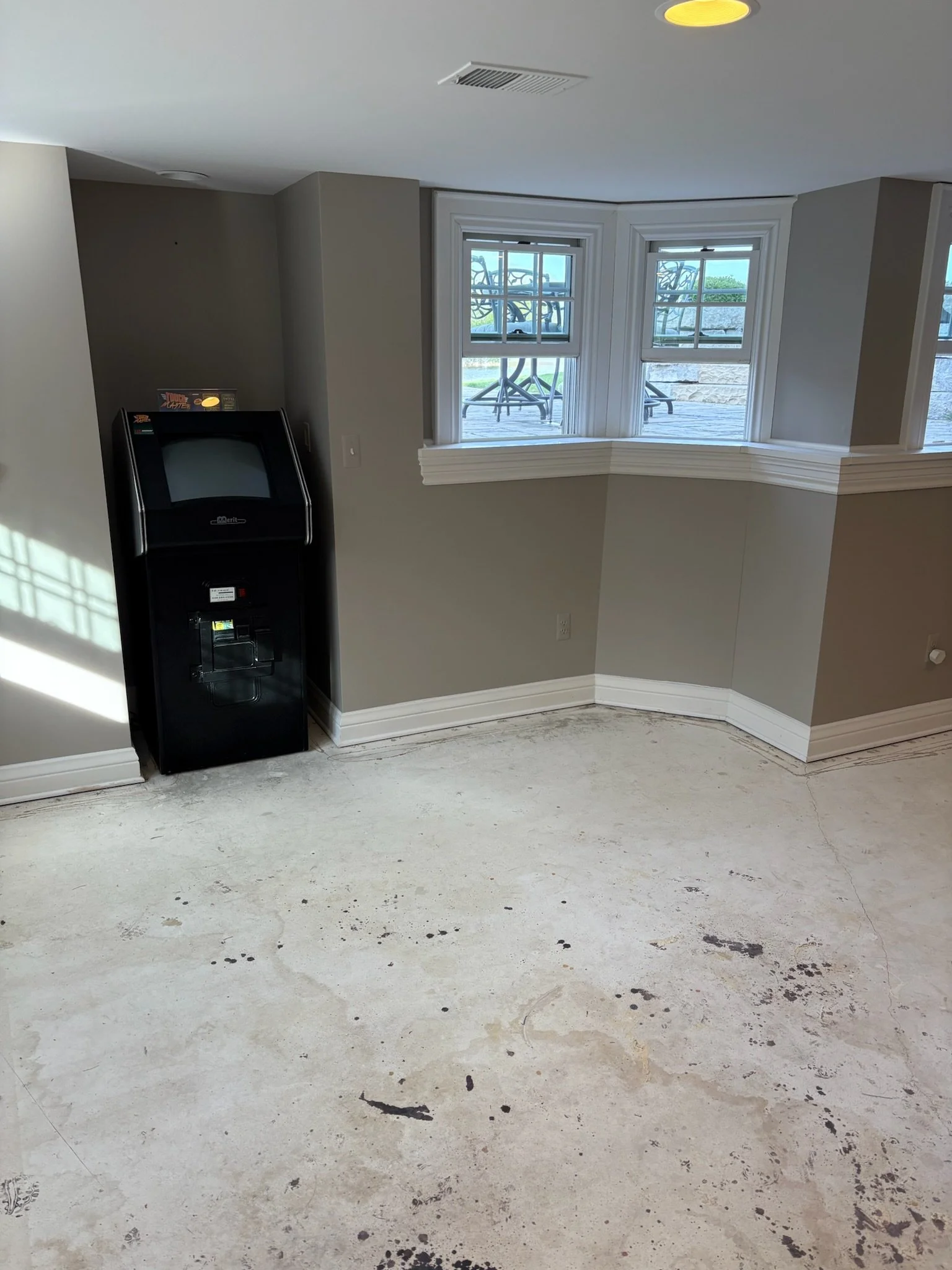 Empty room with unfinished concrete floor, gray walls, a water dispenser, and three windows overlooking an outdoor patio with chairs.