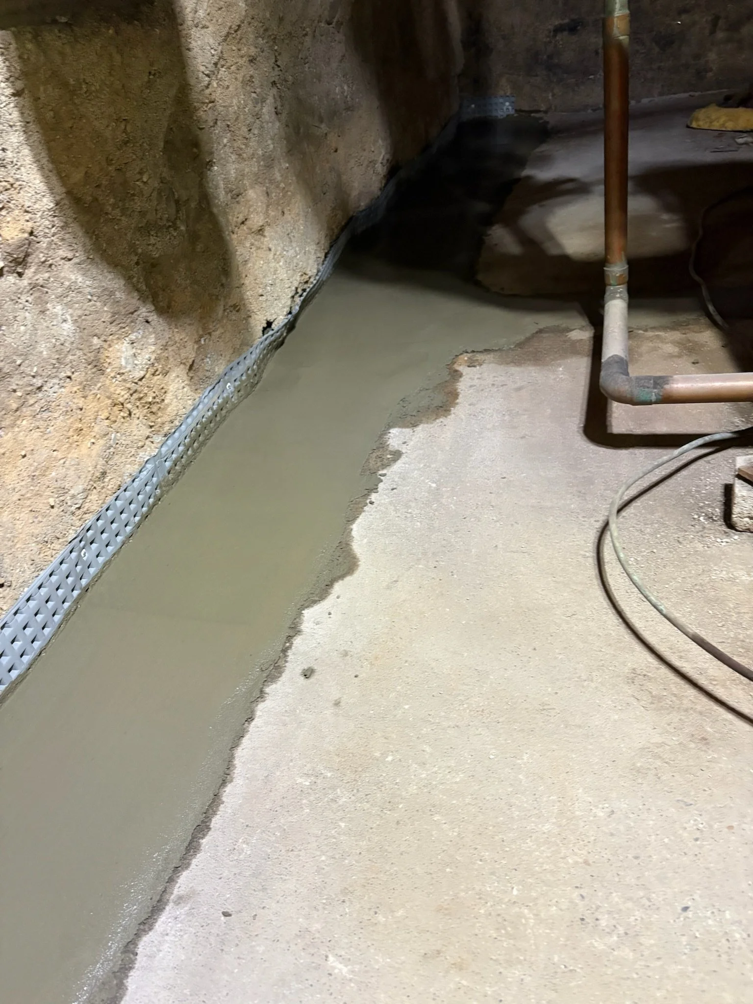 A construction site with fresh concrete poured on the floor, a metal drainage grate along the edge, and exposed insulation and wall materials.