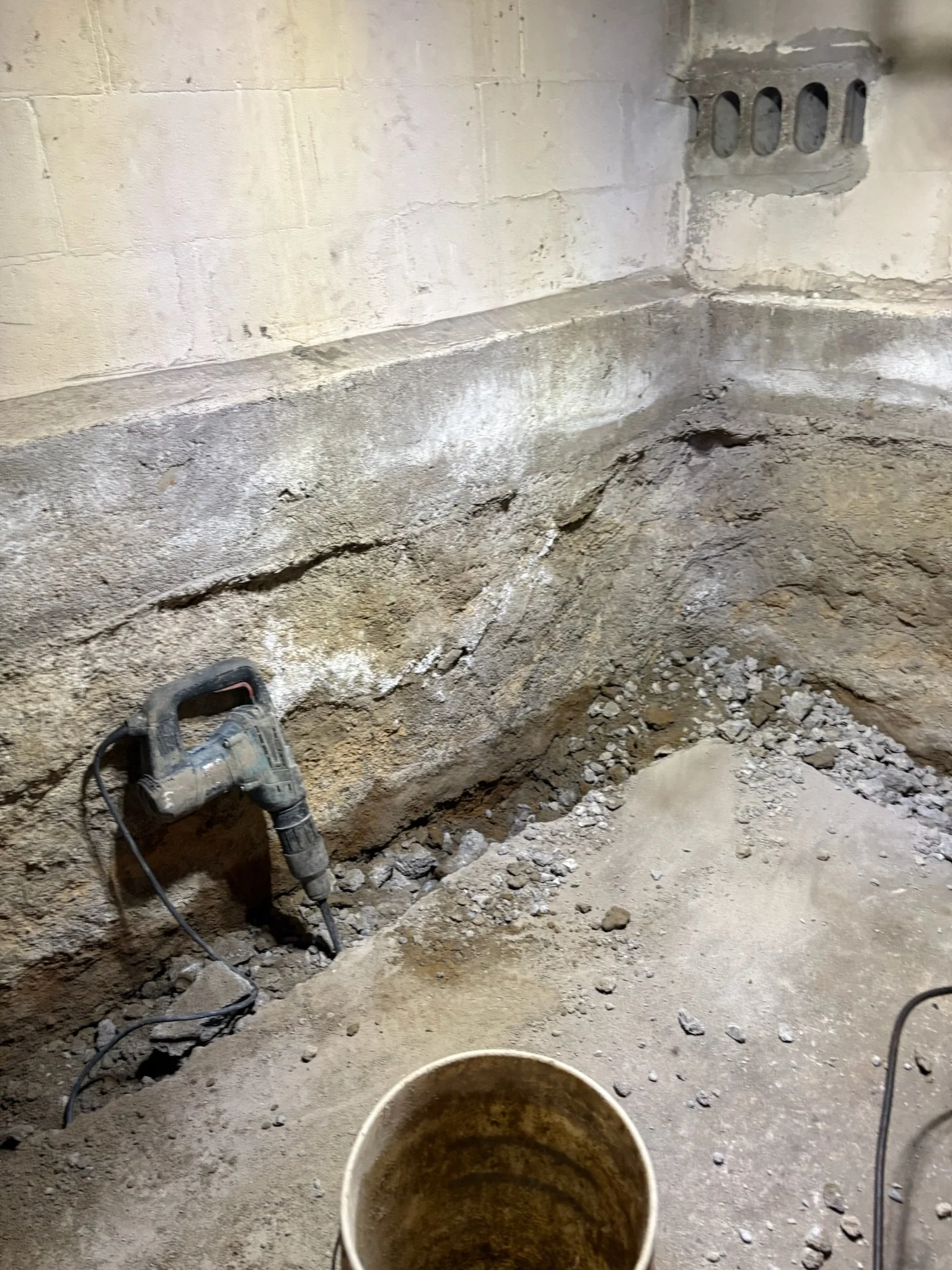 Construction site with exposed soil and rocks, a power drill lying on the ground, and a white bucket in the foreground, with a wall featuring a small vent in the background.
