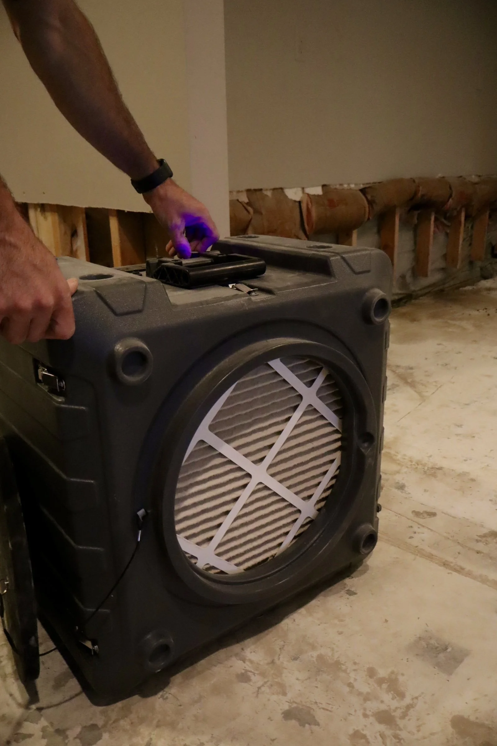 A person moving a portable air purifier with a filter in an unfinished room with exposed wall studs and insulation.