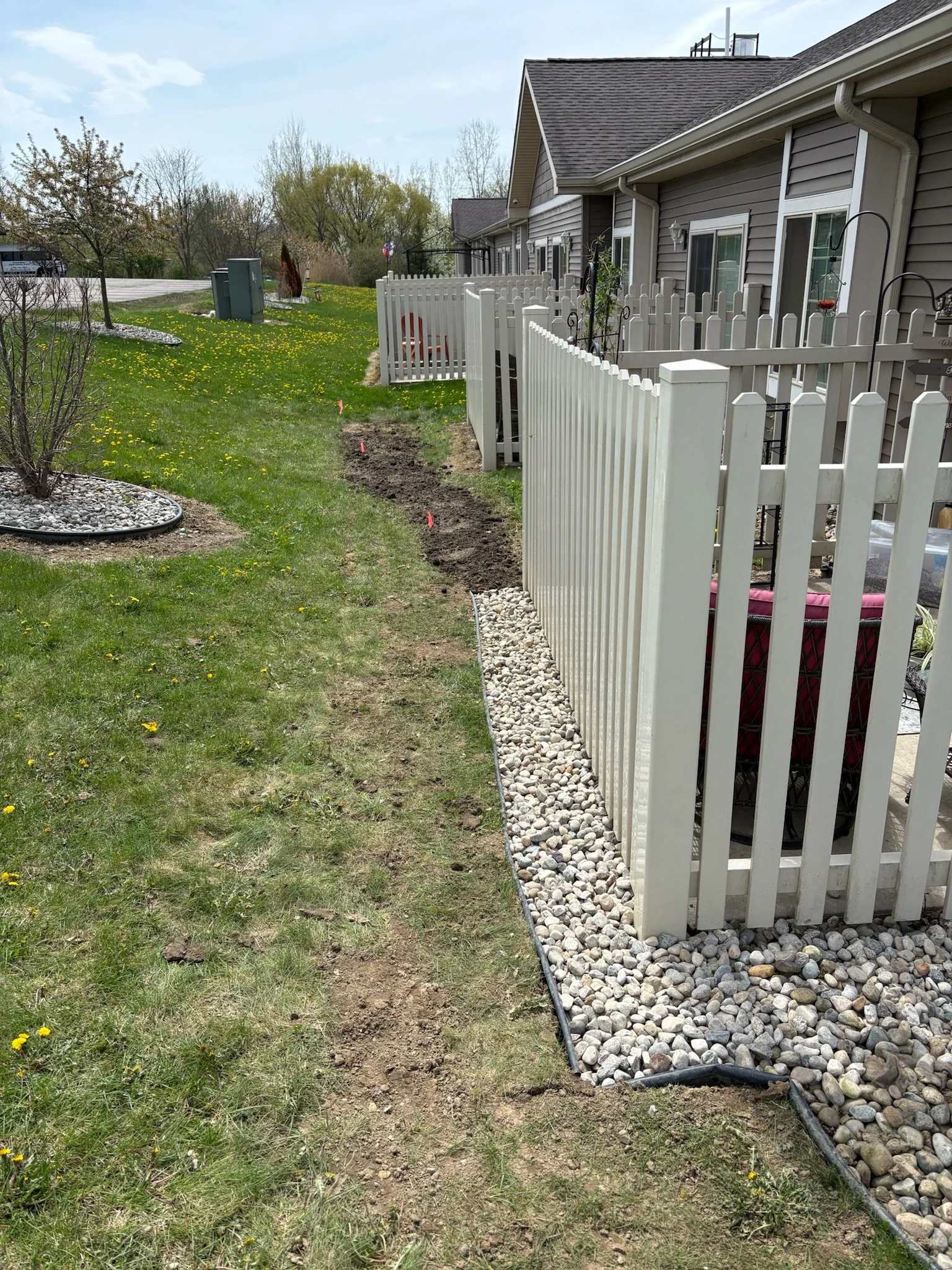 A backyard with a white picket fence, a concrete patio, and a row of grey townhouses. The yard has small trees and yellow flowers, with landscaping and garden beds. A narrow dirt path runs alongside the fence.