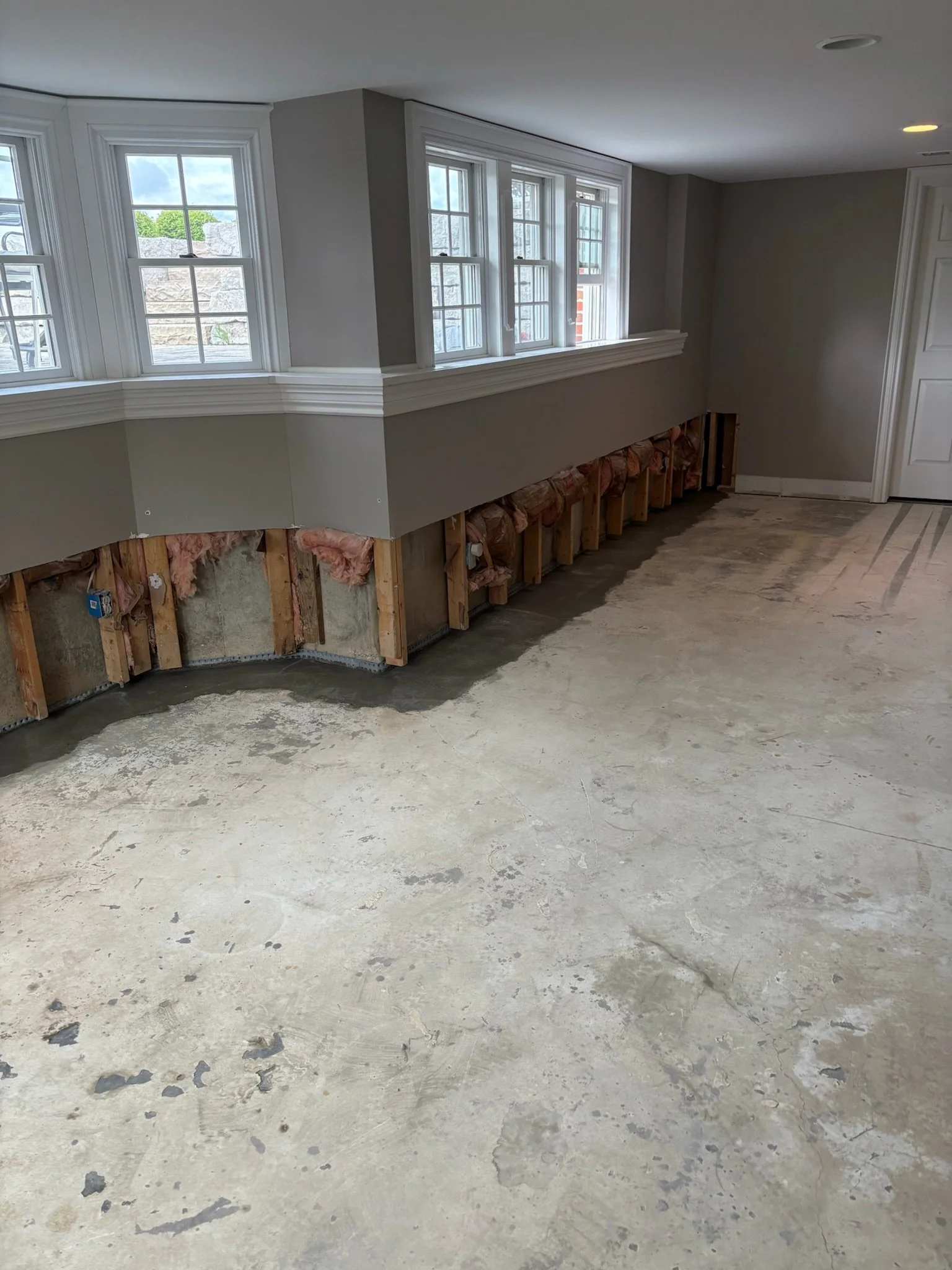Interior of a house under renovation, showing a row of windows, a partially completed wall with insulation and framing, and a bare concrete floor.