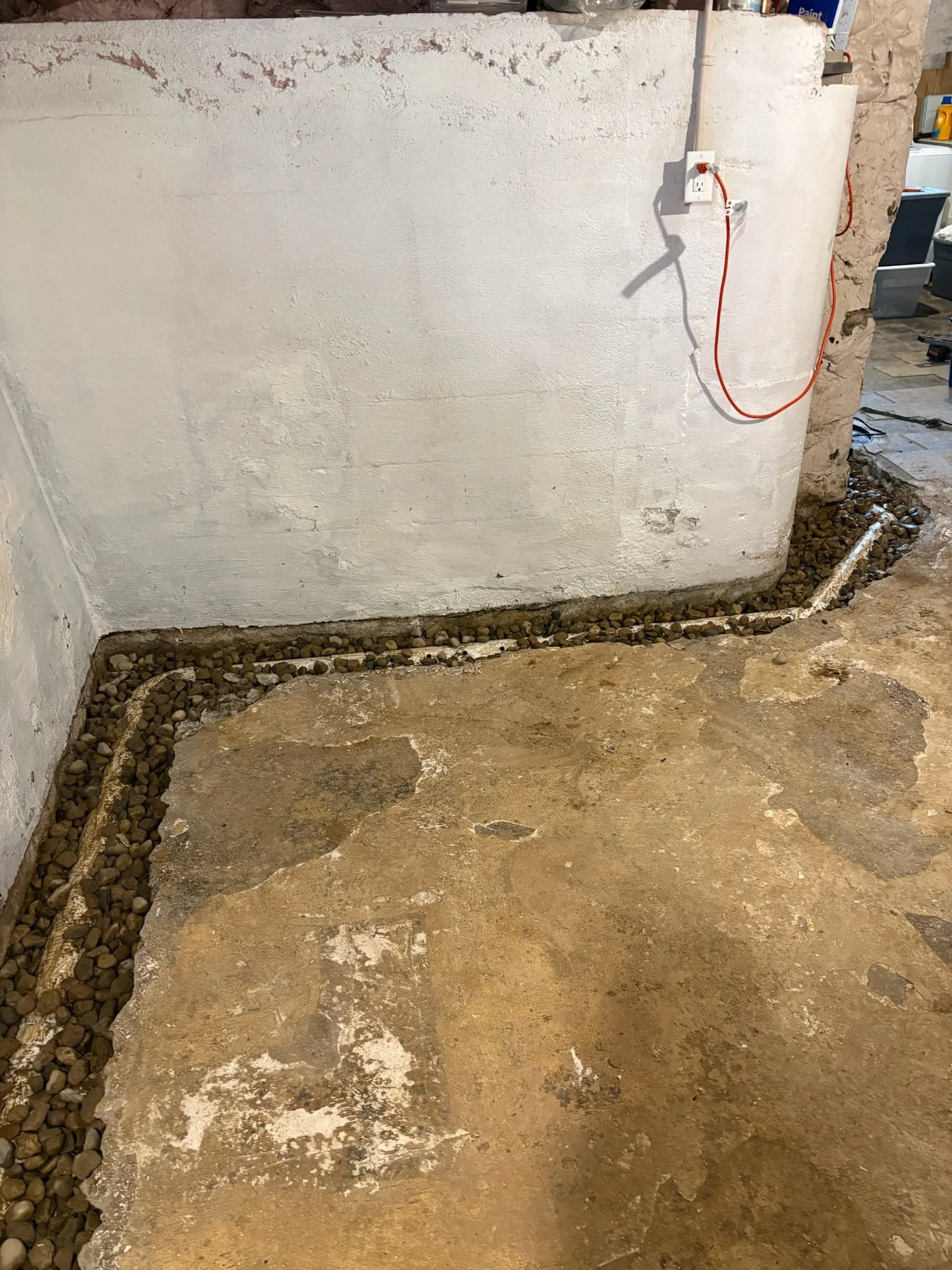 A construction or renovation site showing a partially finished concrete floor, a white wall with some exposed rough patches, and a section of small rocks and gravel along the edge of the flooring. There is also an electrical outlet with a red extensi