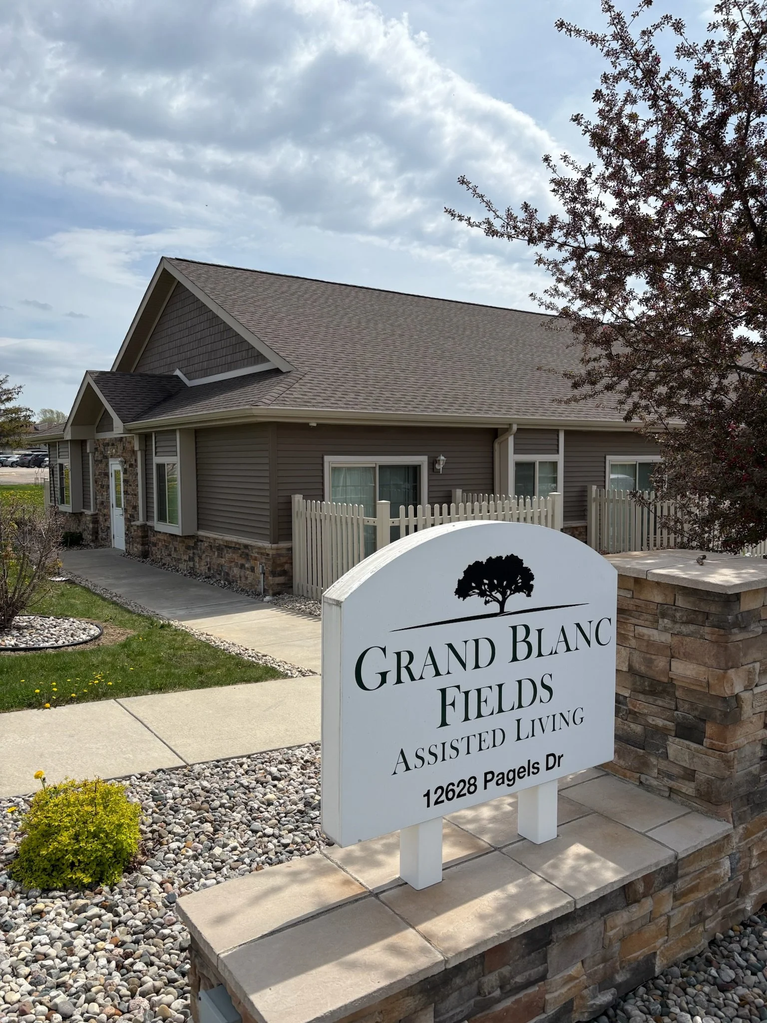 Front view of a residential building with a stone and siding exterior, a paved walkway, a fenced yard, and a sign that reads "Grand Blanc Fields Assisted Living."