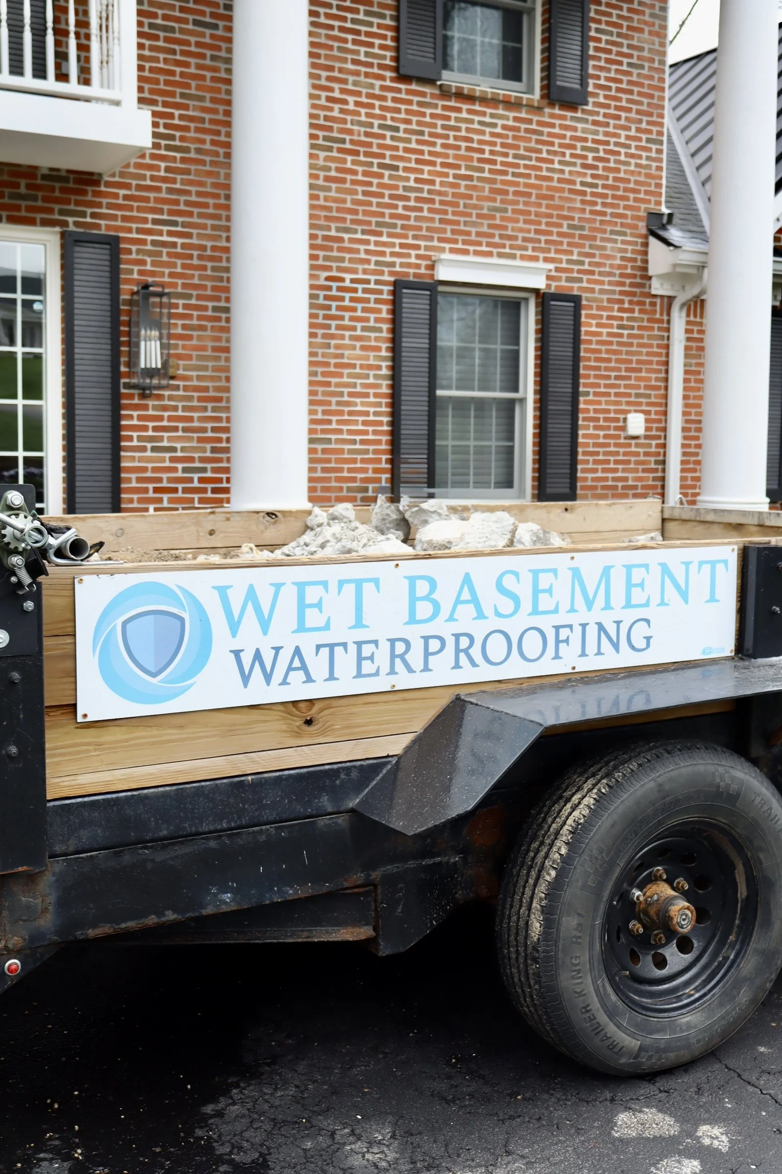 Trailer with a sign reading 'Wet Basement Waterproofing' parked on a driveway in front of a brick house with black window shutters and white columns.