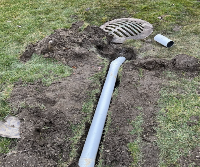 Drainage pipe installation in a grassy yard with dug-up dirt, a storm drain cover, and pipe fittings nearby.
