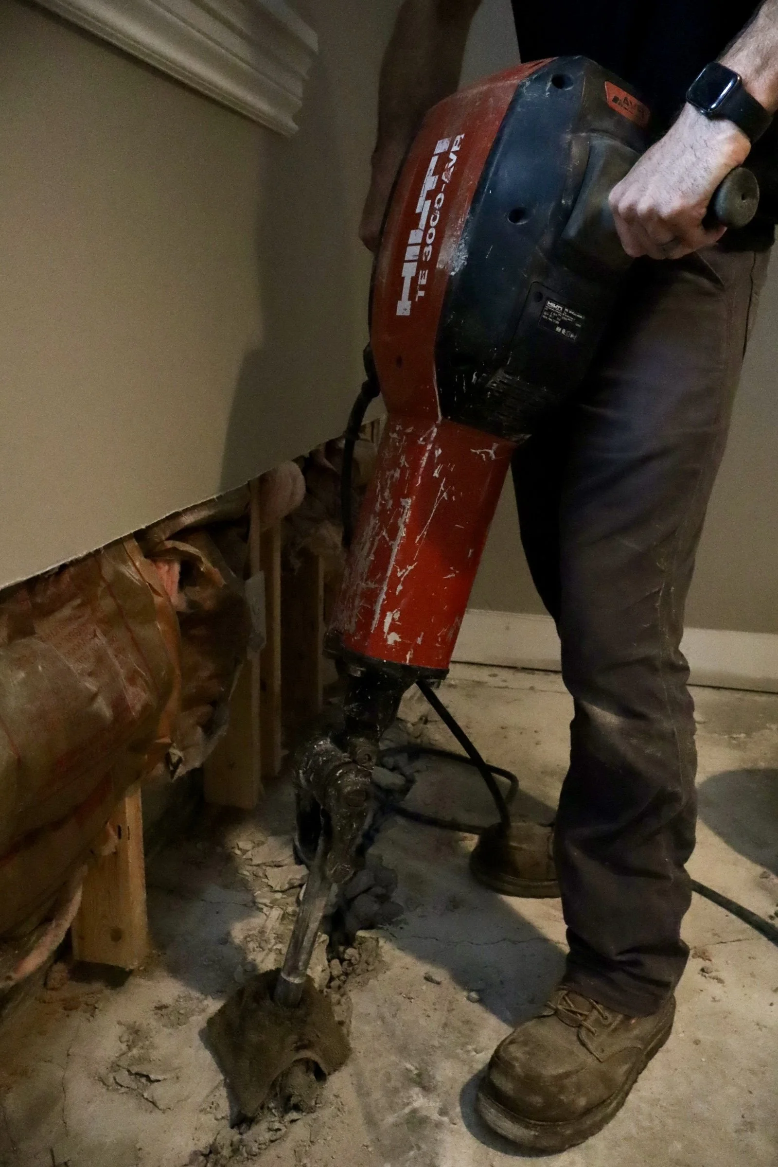 A person wearing leather work boots, brown pants, and a black watch using a red power tool to drill into a wooden wall frame in a room under renovation, with exposed wiring and drywall debris on the floor.