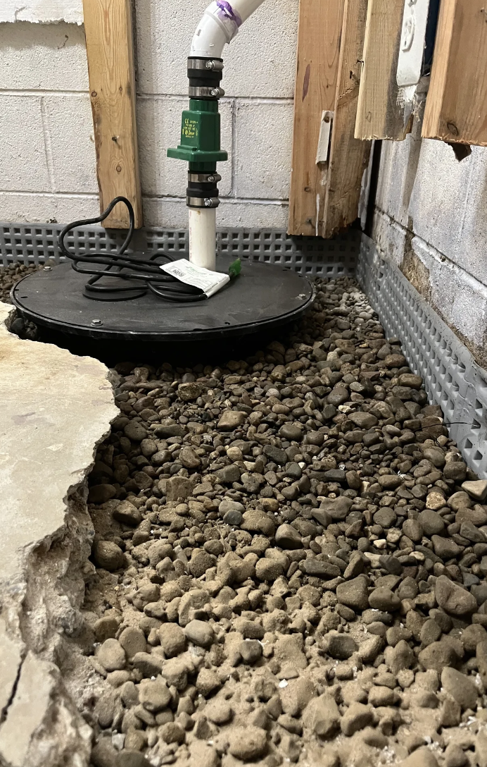 Close-up view of a sump pump setup in a basement or crawl space, with a gravel bed, a plastic cover, a PVC pipe running up, and plumbing connected to the sump pump.
