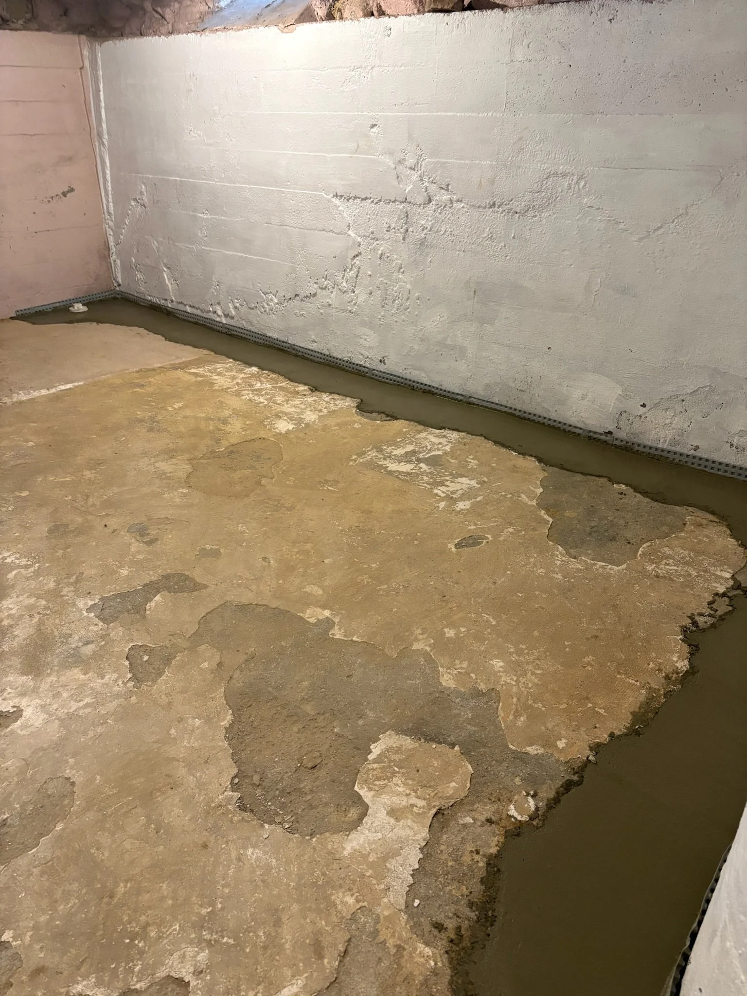Concrete floor with patches of unfinished and damaged areas, with a base of a wall and visible metal channels at the bottom.