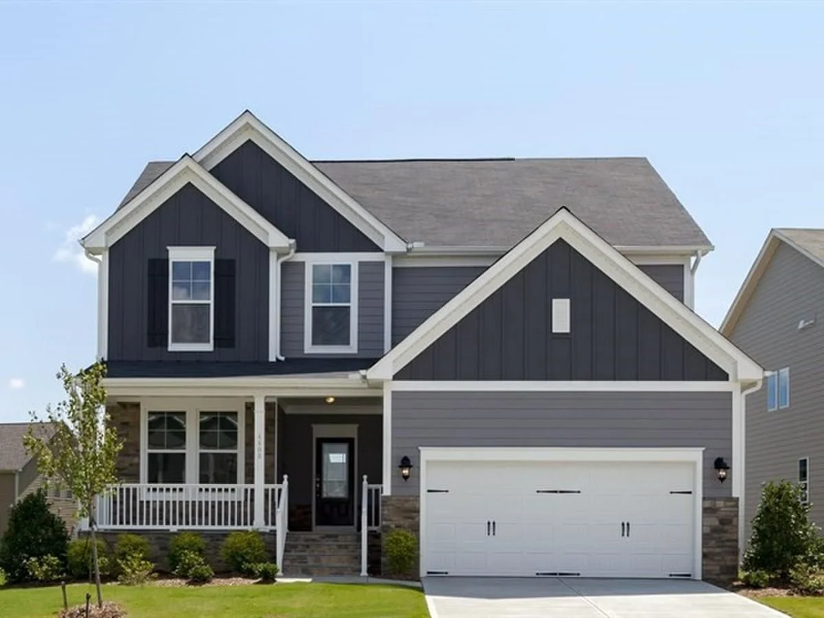 A two-story modern house with gray and black exterior, white trim, a front porch with railings, and an attached two-car garage. The house has a lawn with small shrubs and a young tree in the yard.