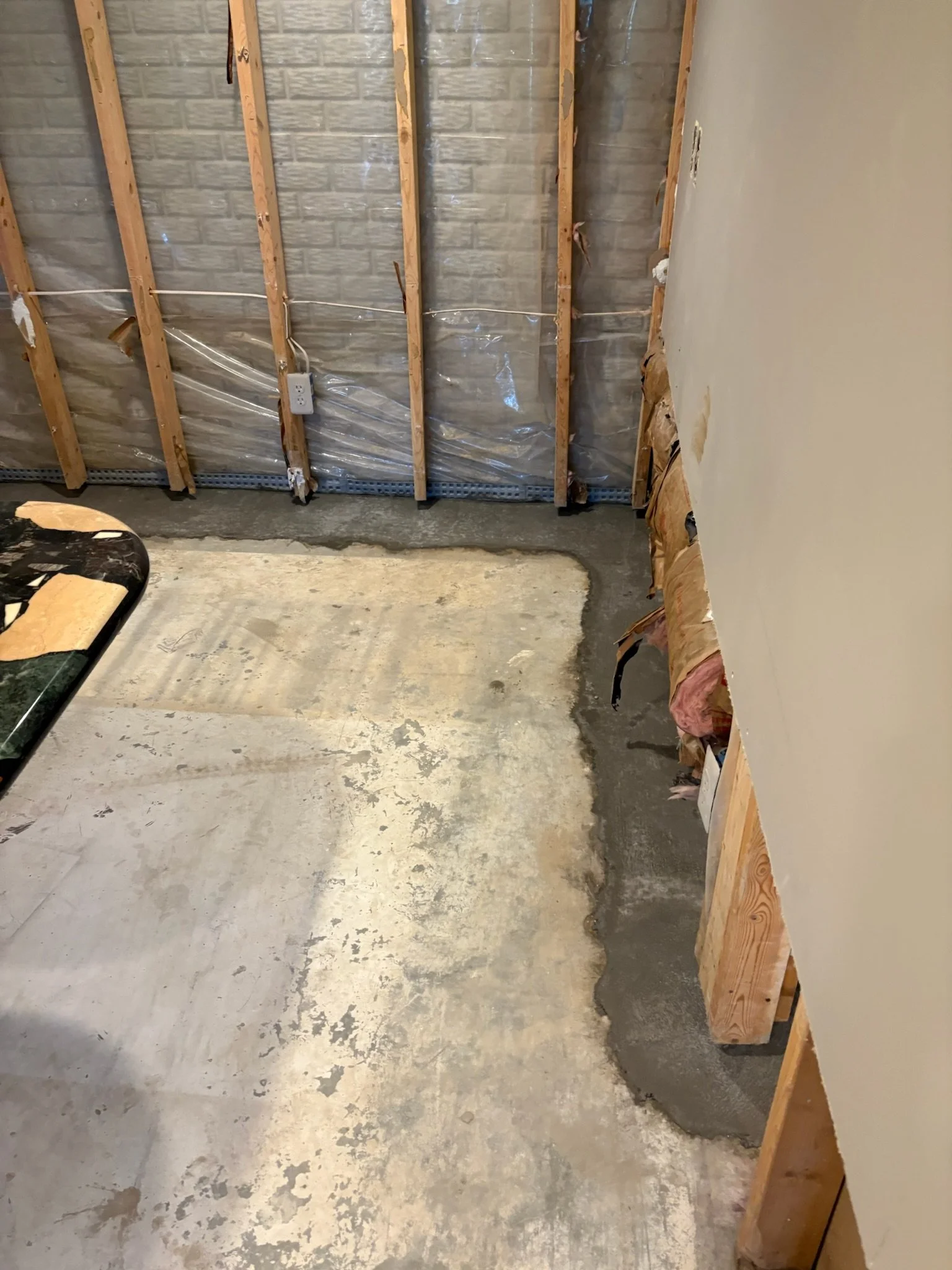 The image shows a wall with exposed wooden studs and electrical wiring, with part of the wall covered by drywall on the right side. The floor is concrete with patches of discoloration.