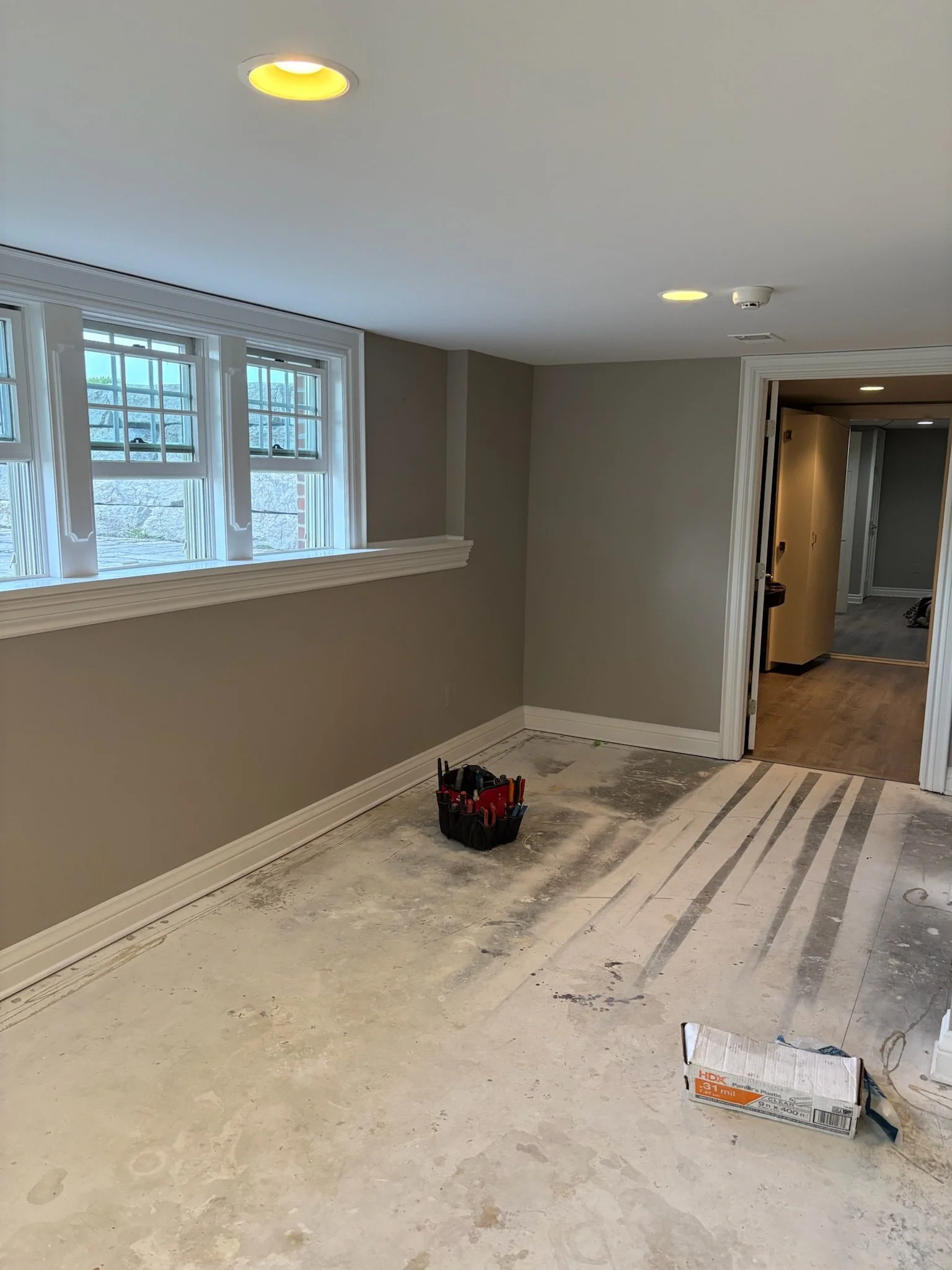 Interior room under renovation with a partially finished floor, a tool bag near the wall, large windows, and a hallway leading to other rooms.