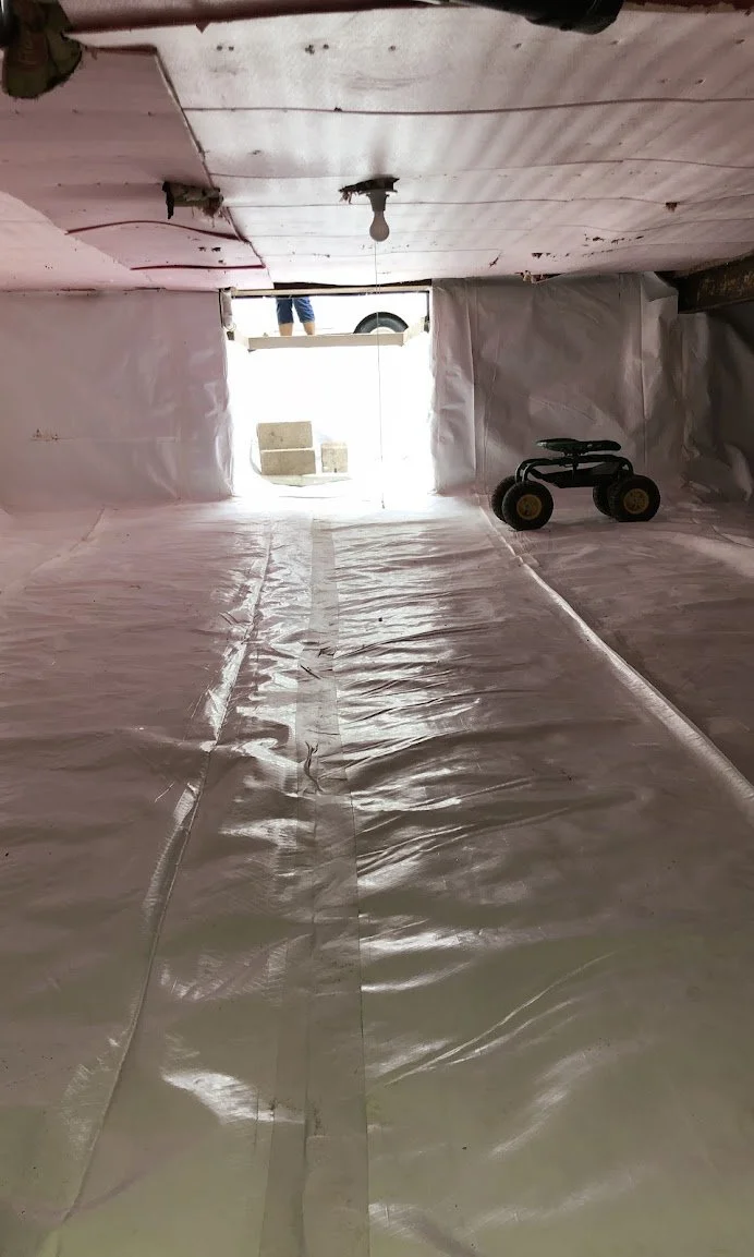 View from inside a construction or renovation site with pink insulation or protective sheeting, a small black and yellow toy vehicle, and a person’s legs visible through an open window or doorway with a bright outdoor scene.