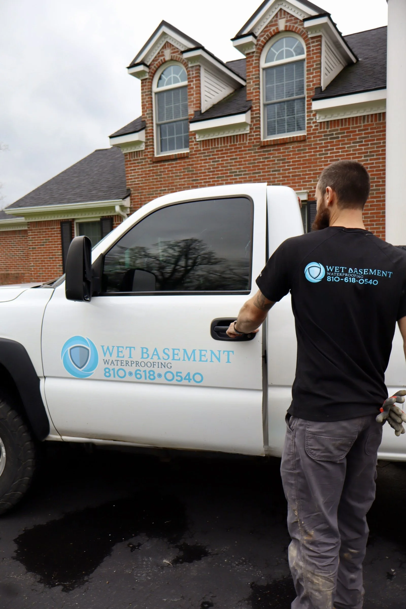 Man wearing a black Wet Basement Waterproofing shirt standing next to a white truck with the company's logo and phone number. The truck is parked in front of a brick house with two large arched windows in the attic and a small window below. The man h