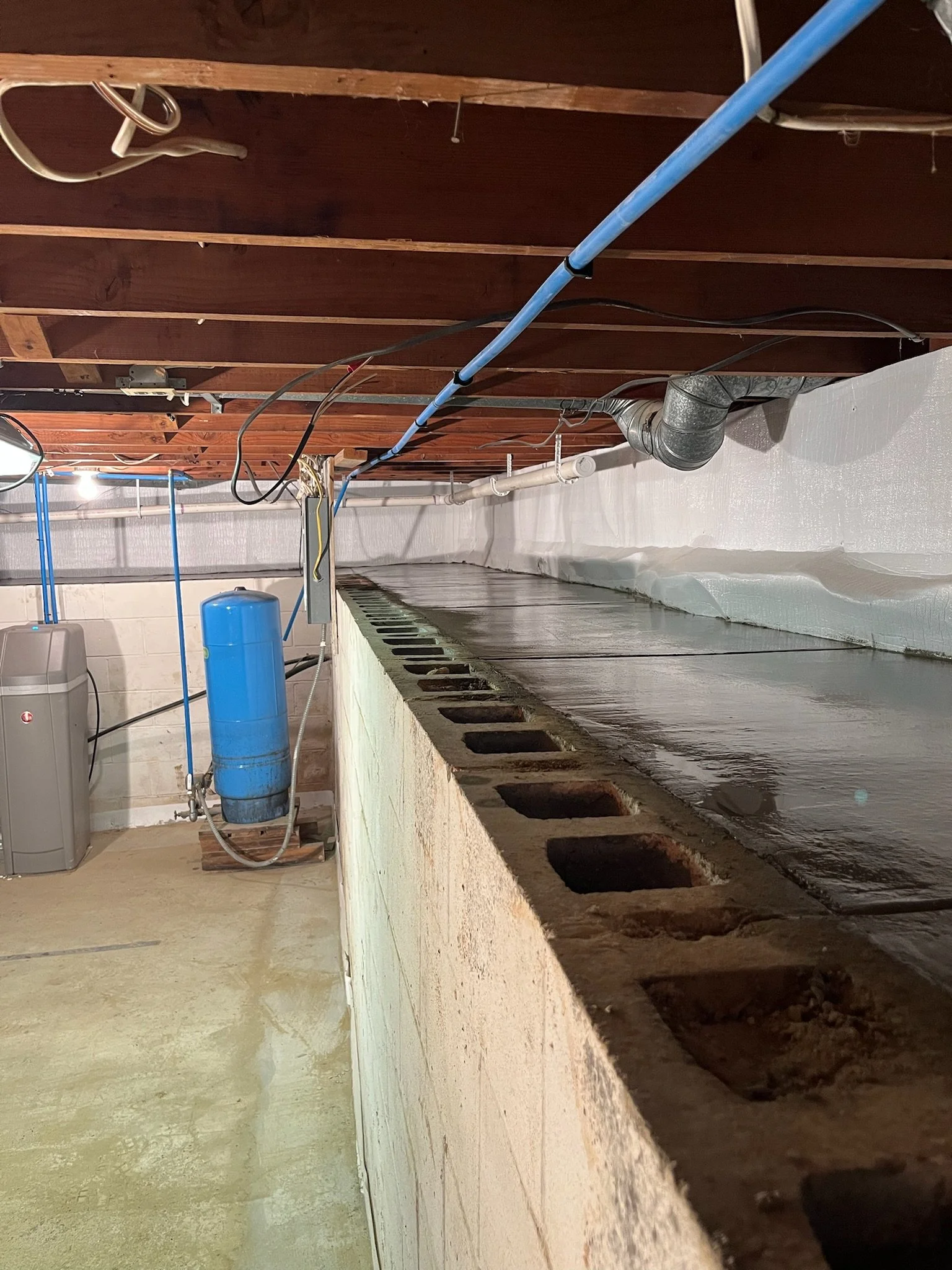 View of a basement with exposed wooden ceiling beams, plumbing pipes, and a concrete floor. There is a blue water pressure tank and a gray water heater in the corner.