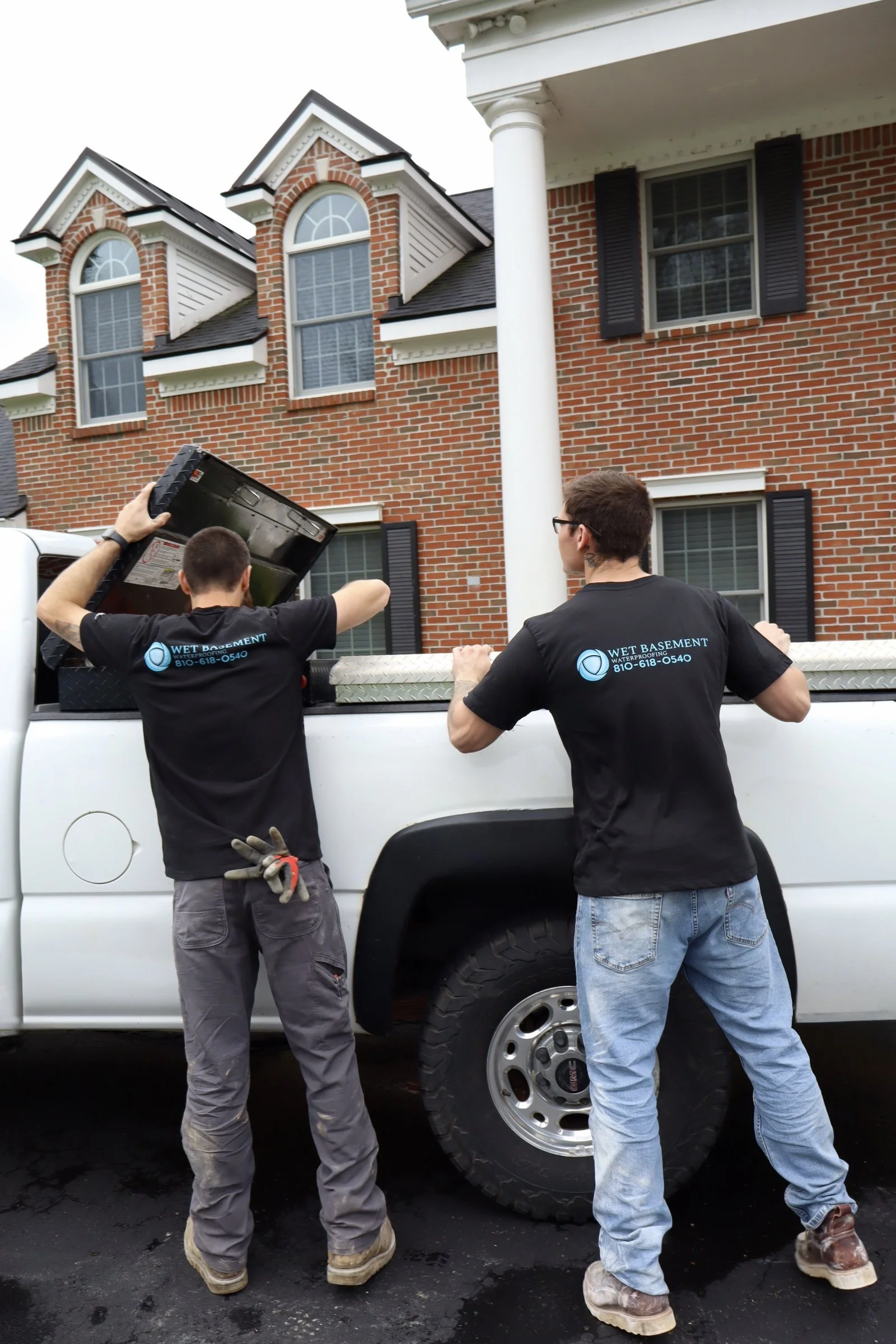 Two men working on a truck, one is lifting an object from the truck bed, and the other is standing near the truck, both wearing black shirts with a company logo and contact number. The truck is parked in front of a brick house.