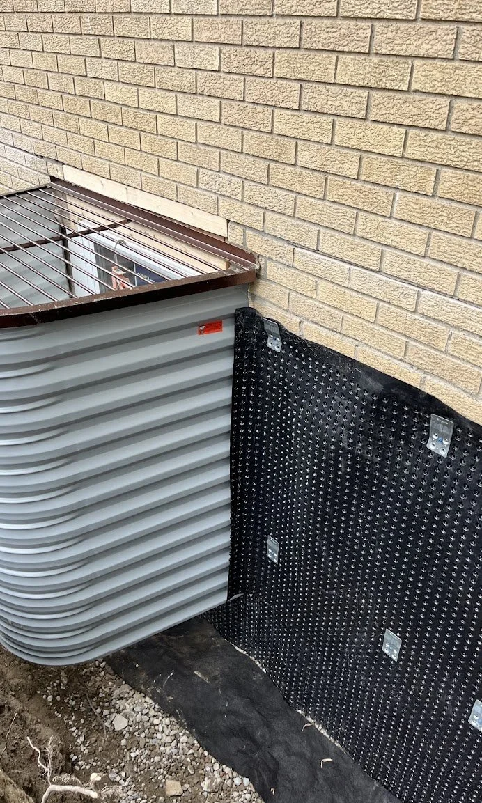 Close-up of an HVAC unit installation next to a beige brick wall, with black waterproof membrane and metal brackets.