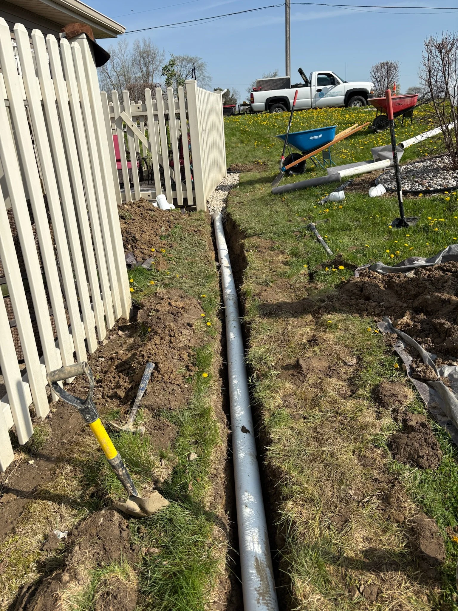 A trench dug in the yard with a white underground pipe laid inside it, surrounded by tools, wheelbarrows, and construction materials, indicating ongoing plumbing or drainage work.