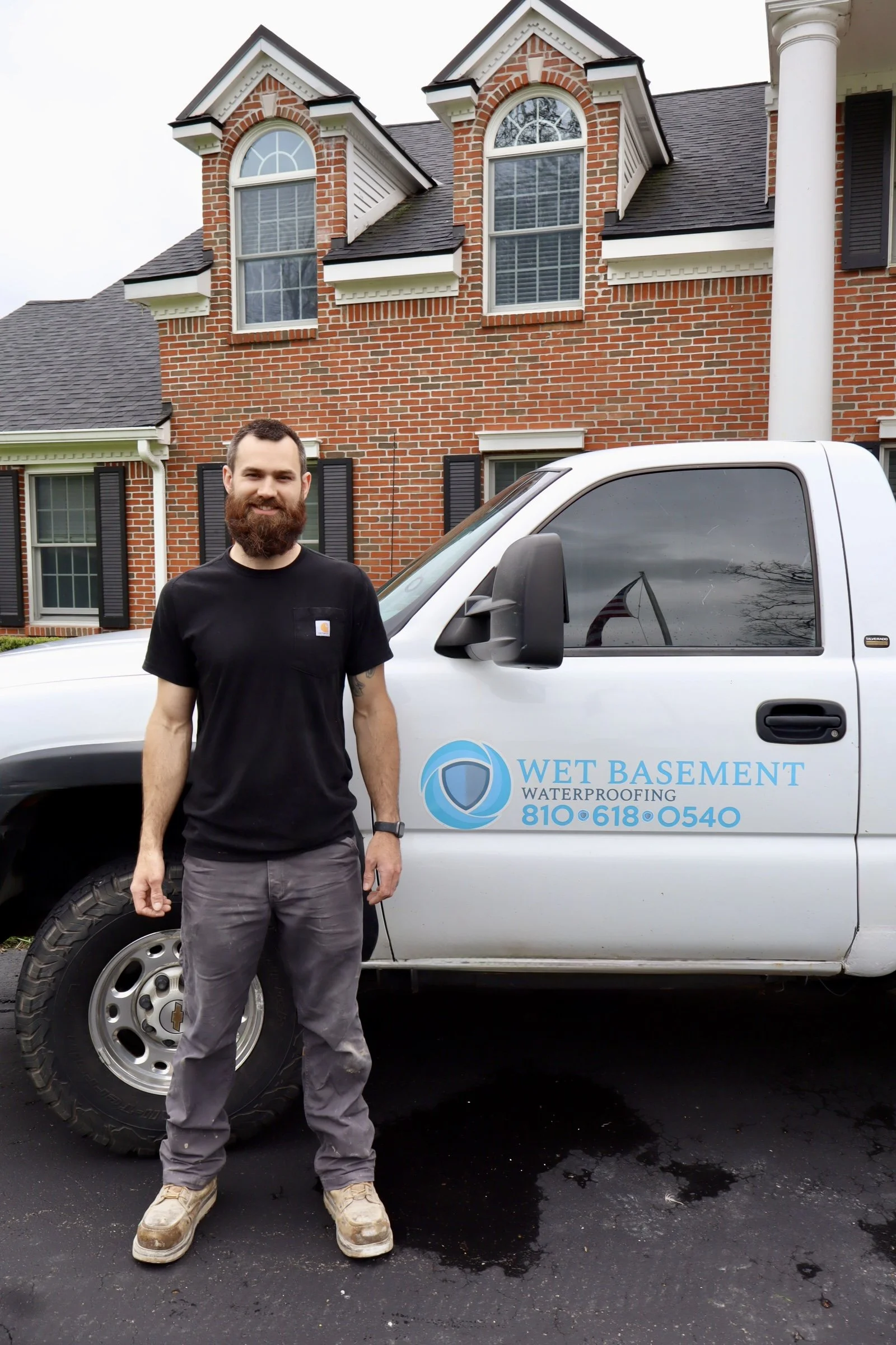 A man with a beard and dark hair standing in front of a white utility truck with the logo and phone number of Wet Basement Waterproofing. The man is wearing a black t-shirt, gray pants, and beige work boots. The background shows a large brick house w