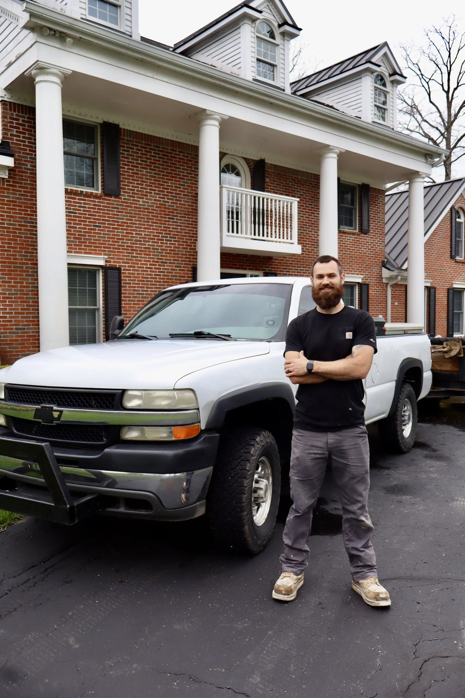 A man with a beard wearing a black t-shirt, gray pants, and sneakers standing in front of a white pickup truck parked on asphalt driveway in front of a brick house with white columns and dormer windows.