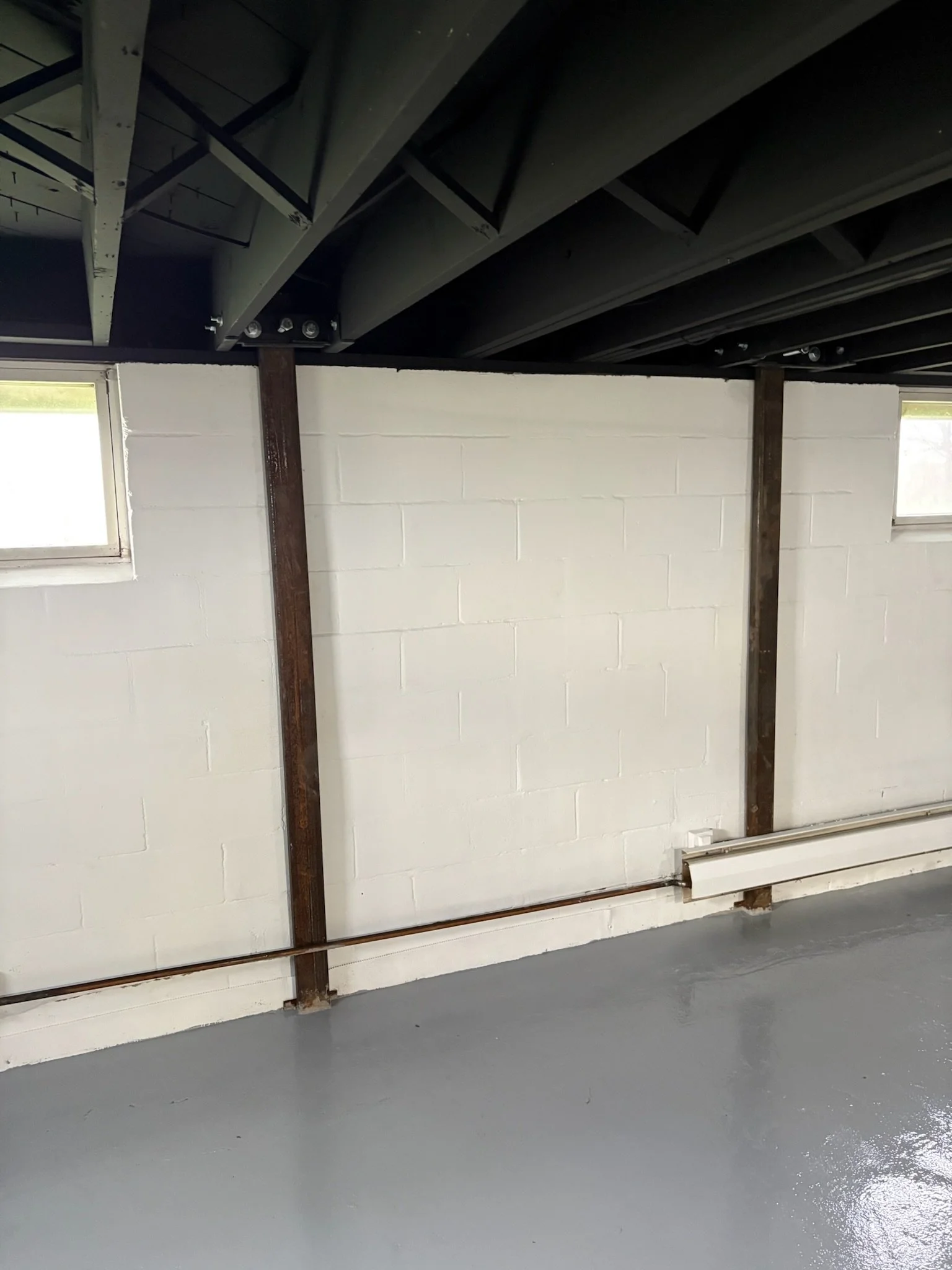 The image shows an unfinished basement with a white cinder block wall, two small windows, and metal support beams. The concrete floor is painted gray and looks freshly coated.
