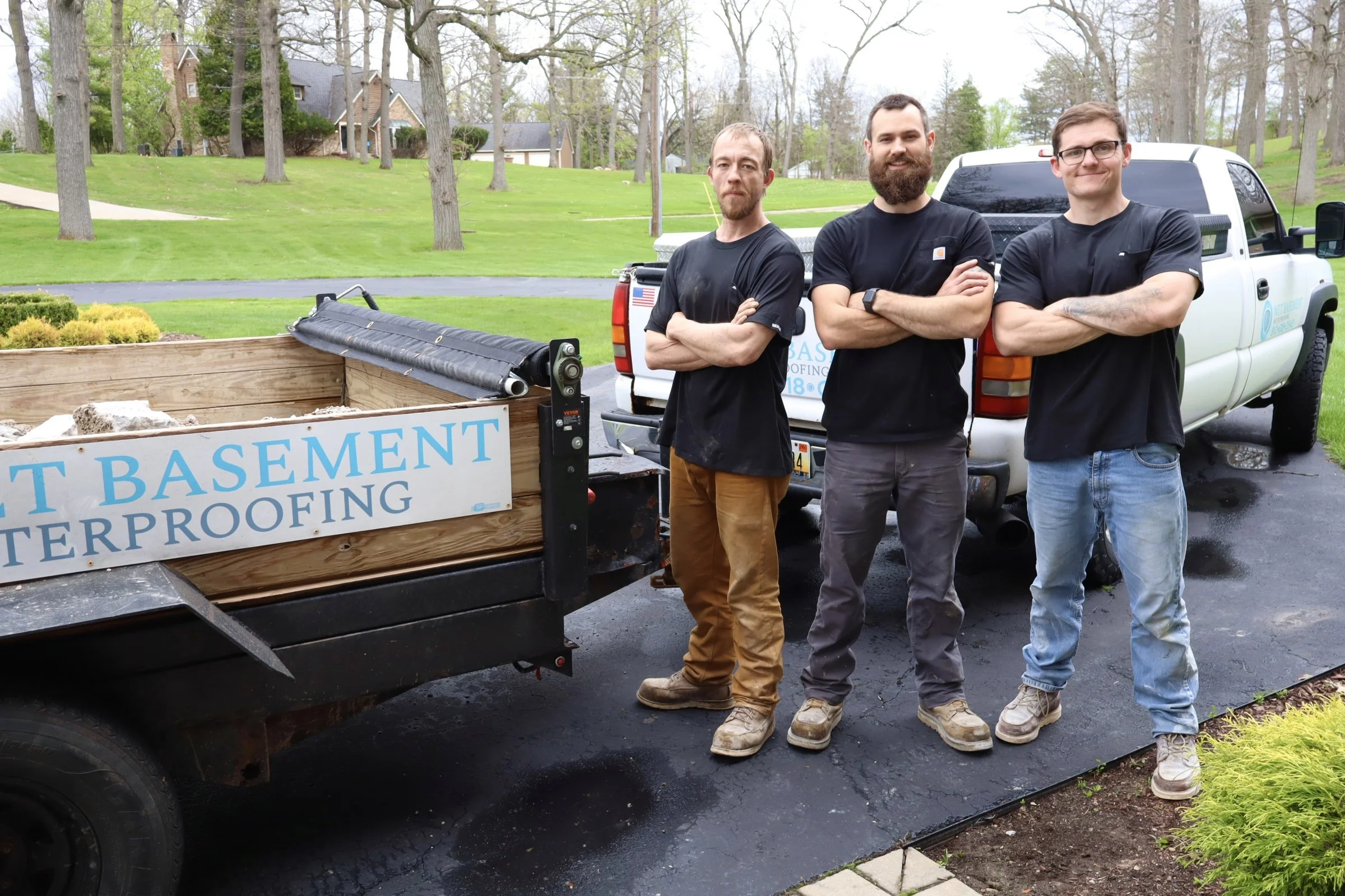 Three men standing with arms crossed in front of a pickup truck, with a sign that reads 'Basement Waterproofing'. They are outdoors on a driveway in a neighborhood with green lawns and trees.