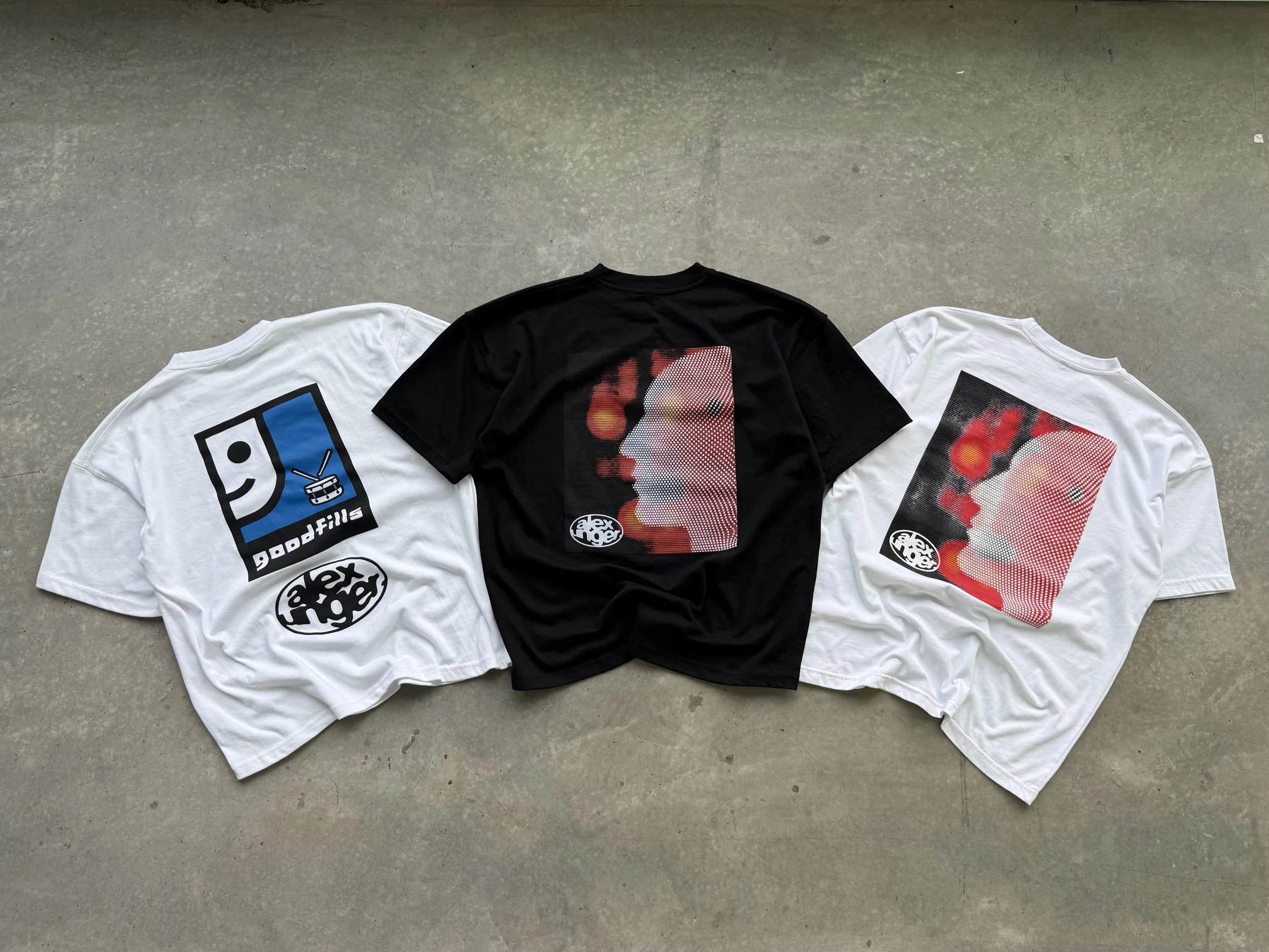 Three t-shirts laid flat on a concrete floor, two white with graphic designs and one black with a different graphic design, featuring faces and logo patterns.