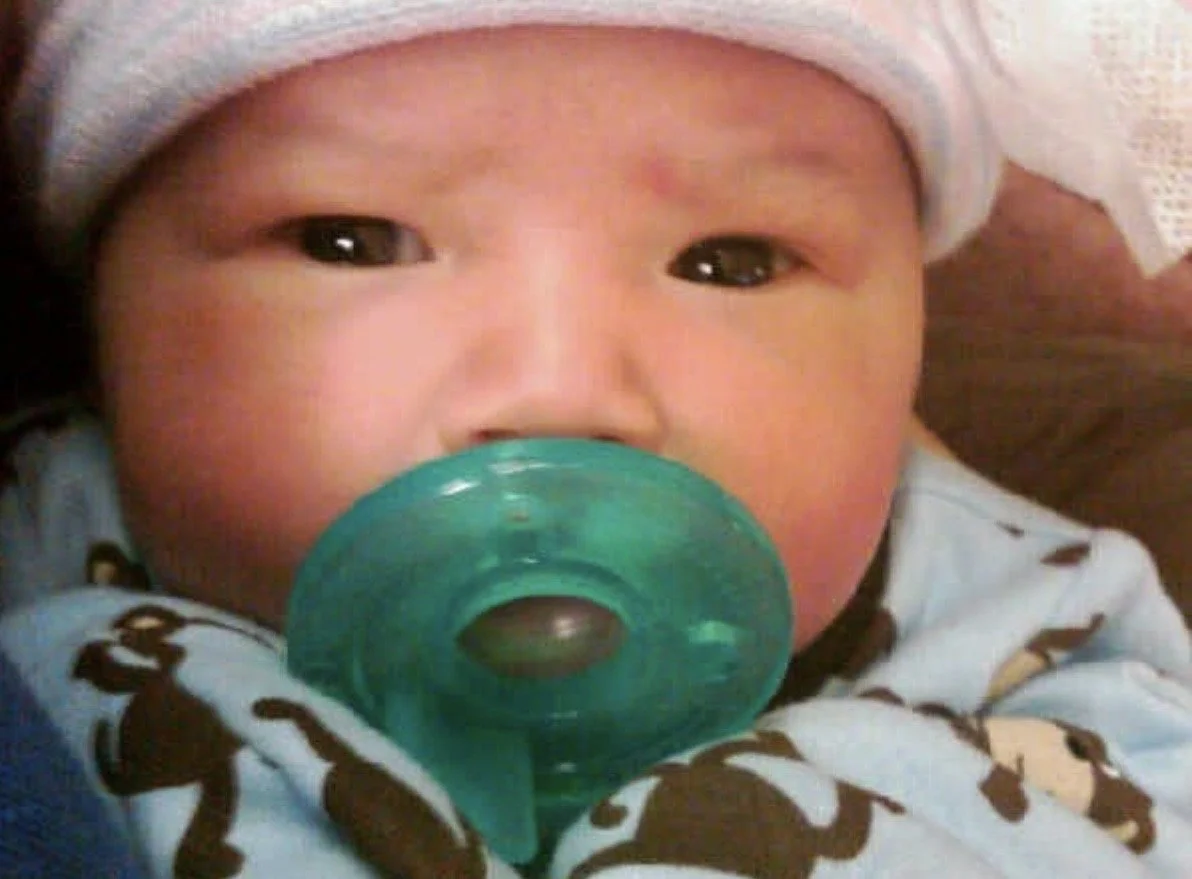 Close-up of a baby with a pacifier in their mouth, wearing a white hat and a garment with black monkey patterns.
