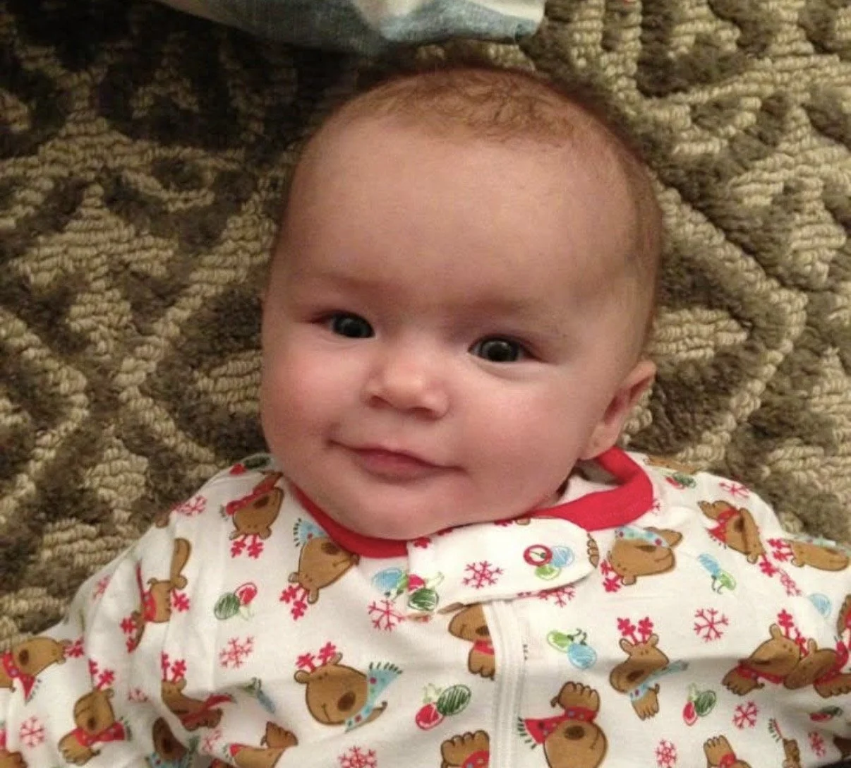 Close-up of a smiling baby with light skin and dark hair, wearing a Christmas-themed onesie with a red collar and reindeer pattern, lying on a textured brown blanket.