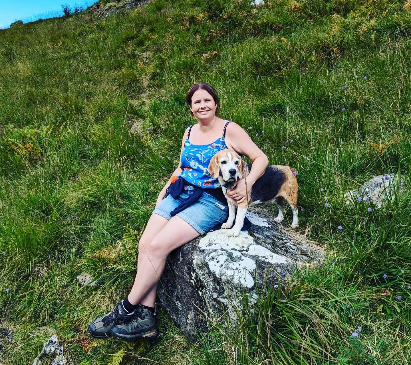 A woman sitting on a large rock with a beagle dog in a grassy, hilly outdoor area.
