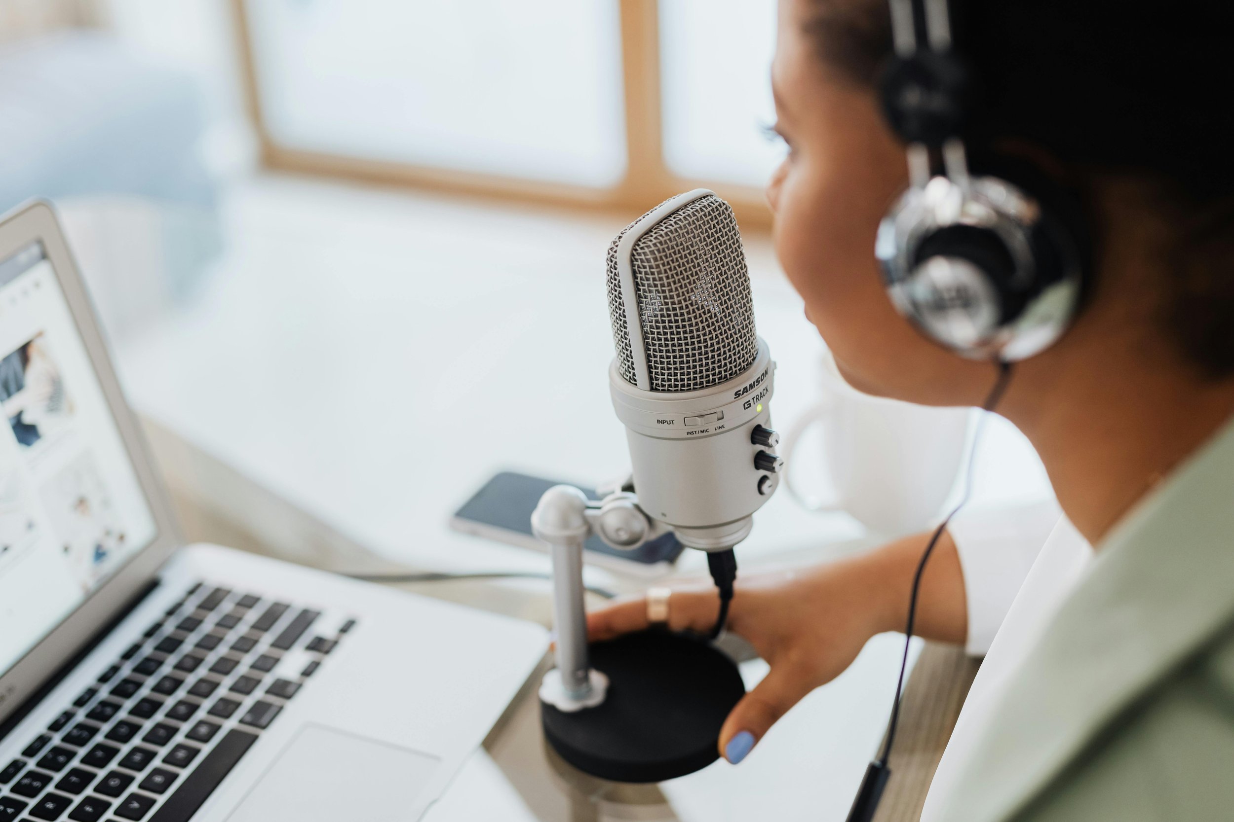 A woman wearing headphones speaking into a professional microphone during a recording session or podcast. There is a laptop and a tablet on the desk in front of her.