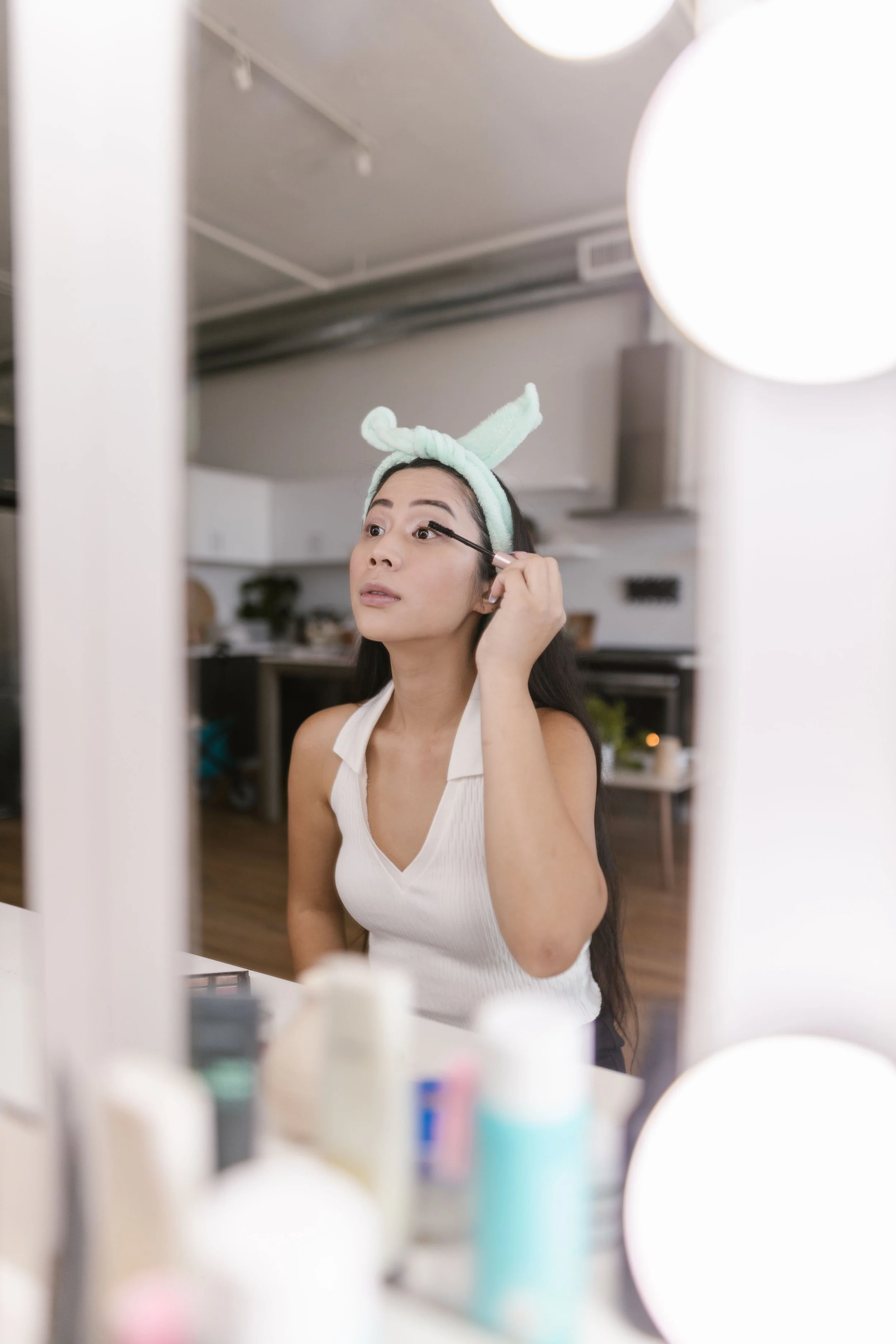 A woman applying mascara to her eyelashes while looking into a mirror in a well-lit room, with bottles and containers visible in the foreground.