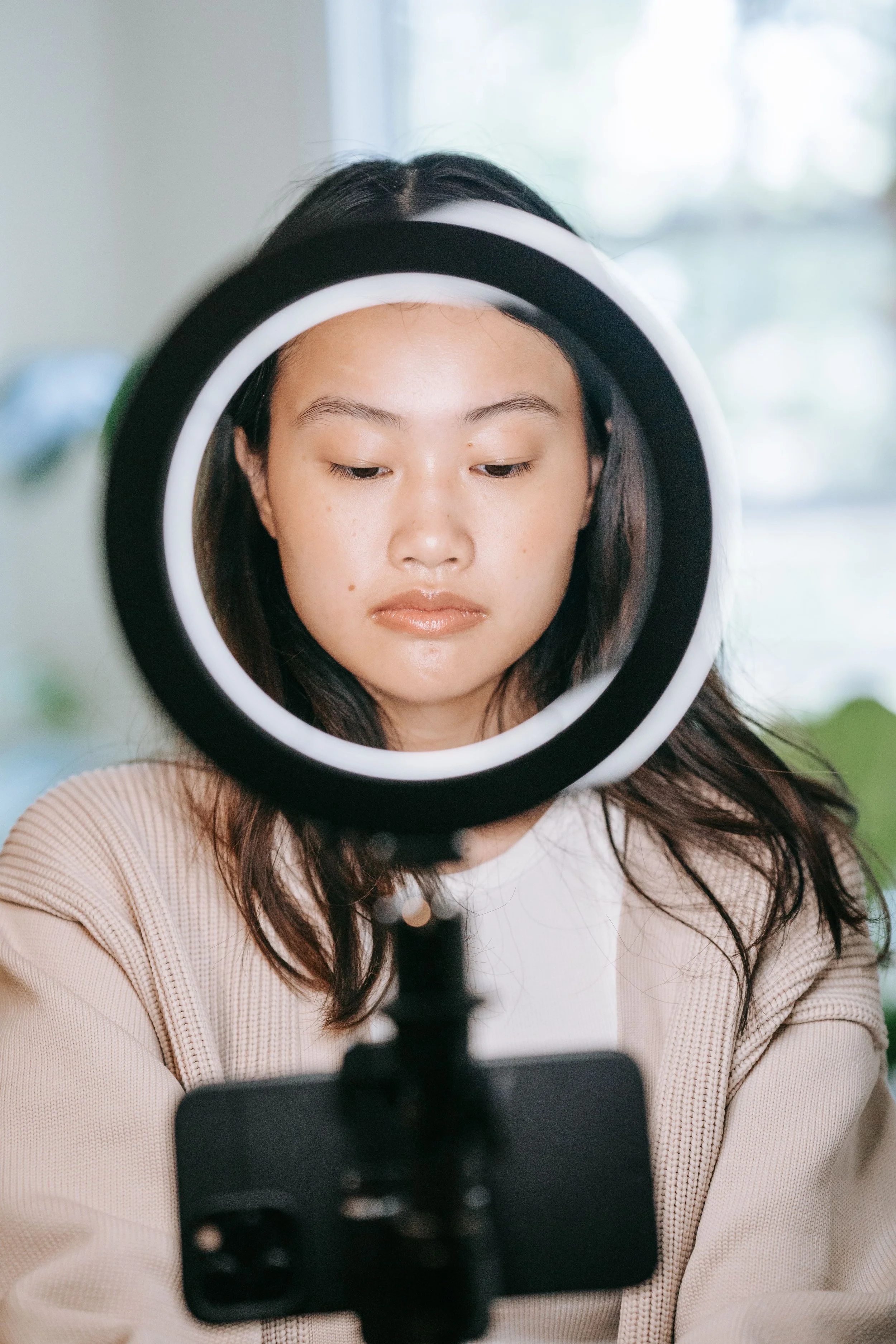 A woman looking down at her reflection in a circular mirror that is mounted on a stand, with a camera or phone set up in front of her to take the photo.