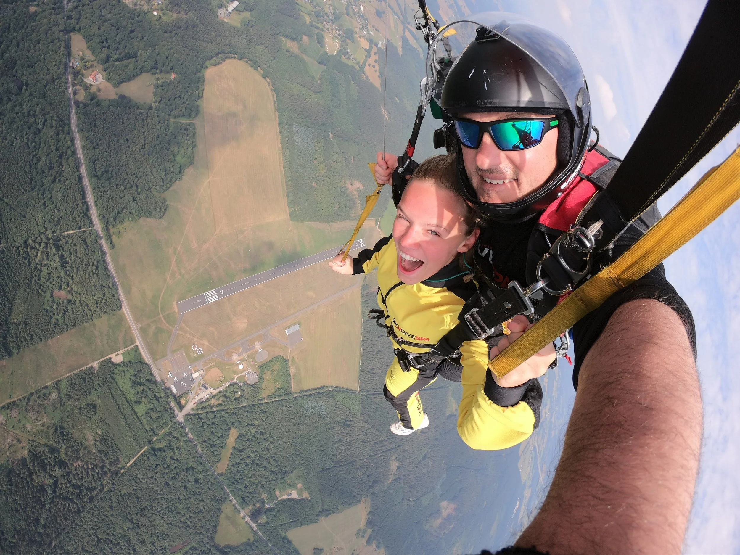 Two people skydiving, with one taking a selfie, high above a landscape of fields, forests, and a runway.