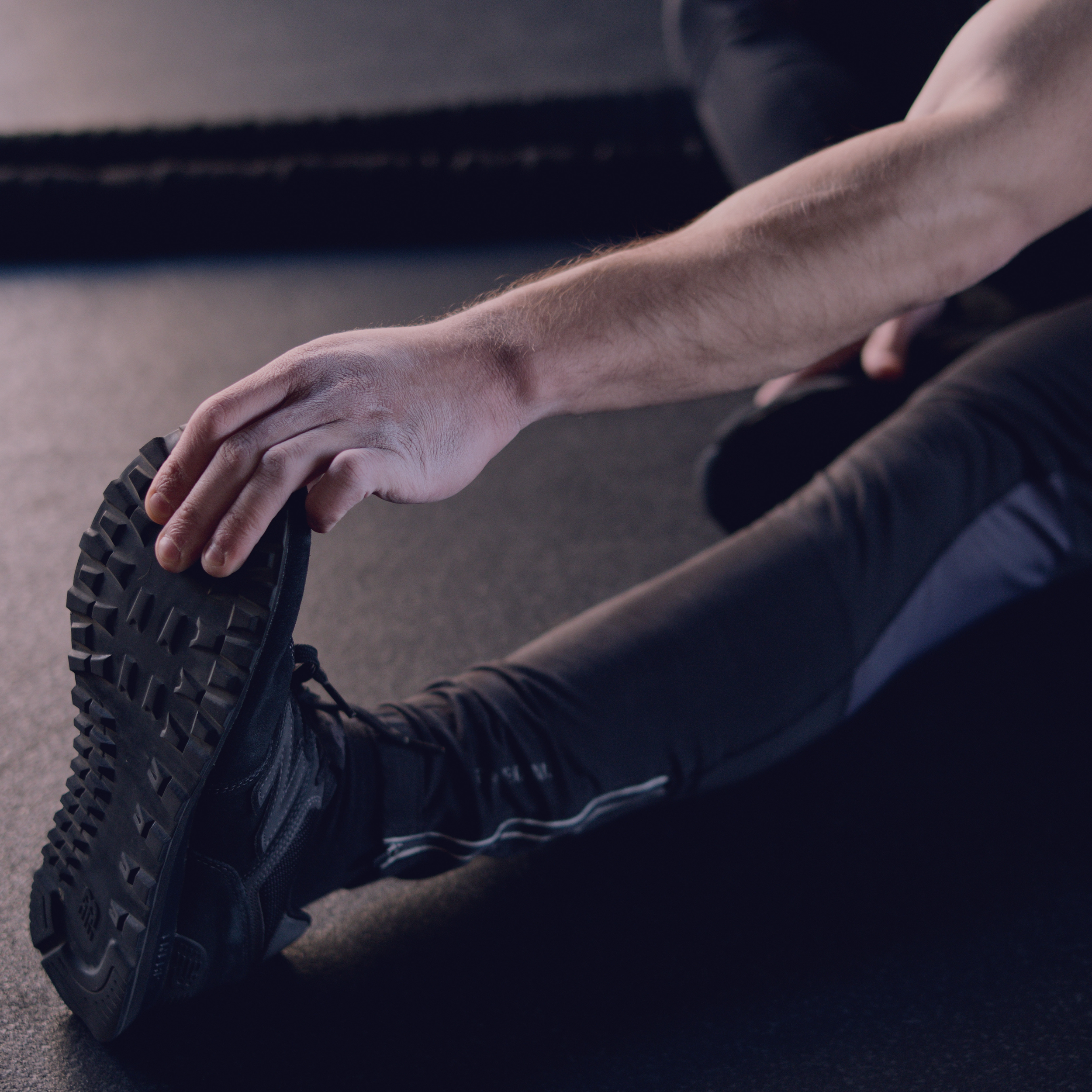 Close-up of a person's bent leg and foot wearing a black sneaker with a rugged tire sole, on a black athletic shoe, on a dark surface.