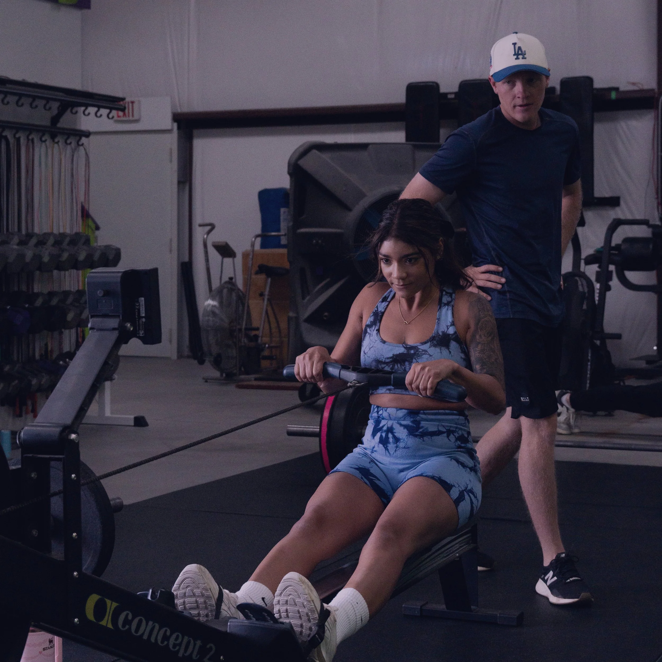 A man wearing a white and blue Los Angeles Dodgers baseball cap and athletic clothing is standing in a gym, with a young woman exercising on a treadmill in the foreground. The gym has weight racks, exercise machines, and bookshelves in the background.