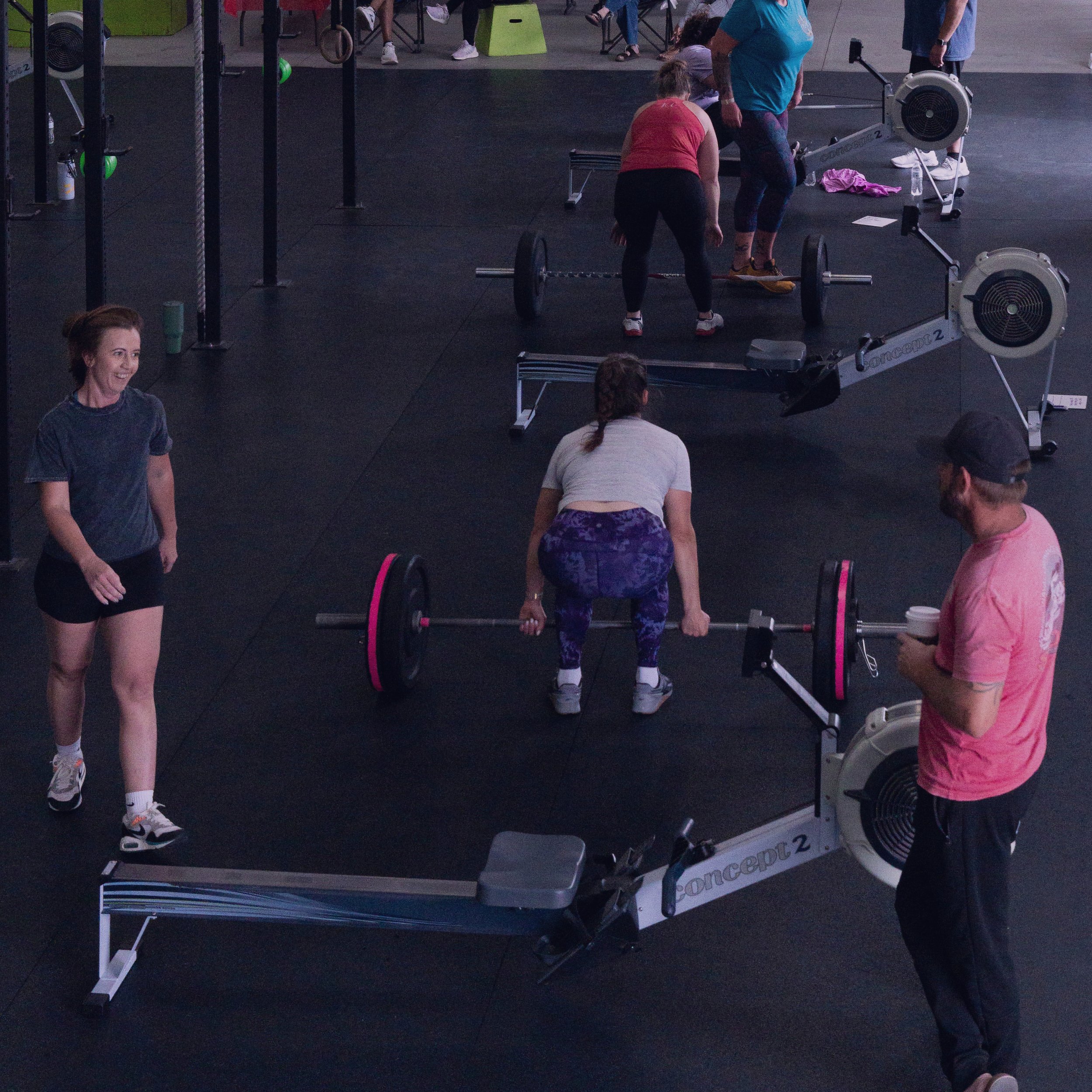 People participating in a CrossFit class, lifting barbells, in a gym with black floors and equipment.