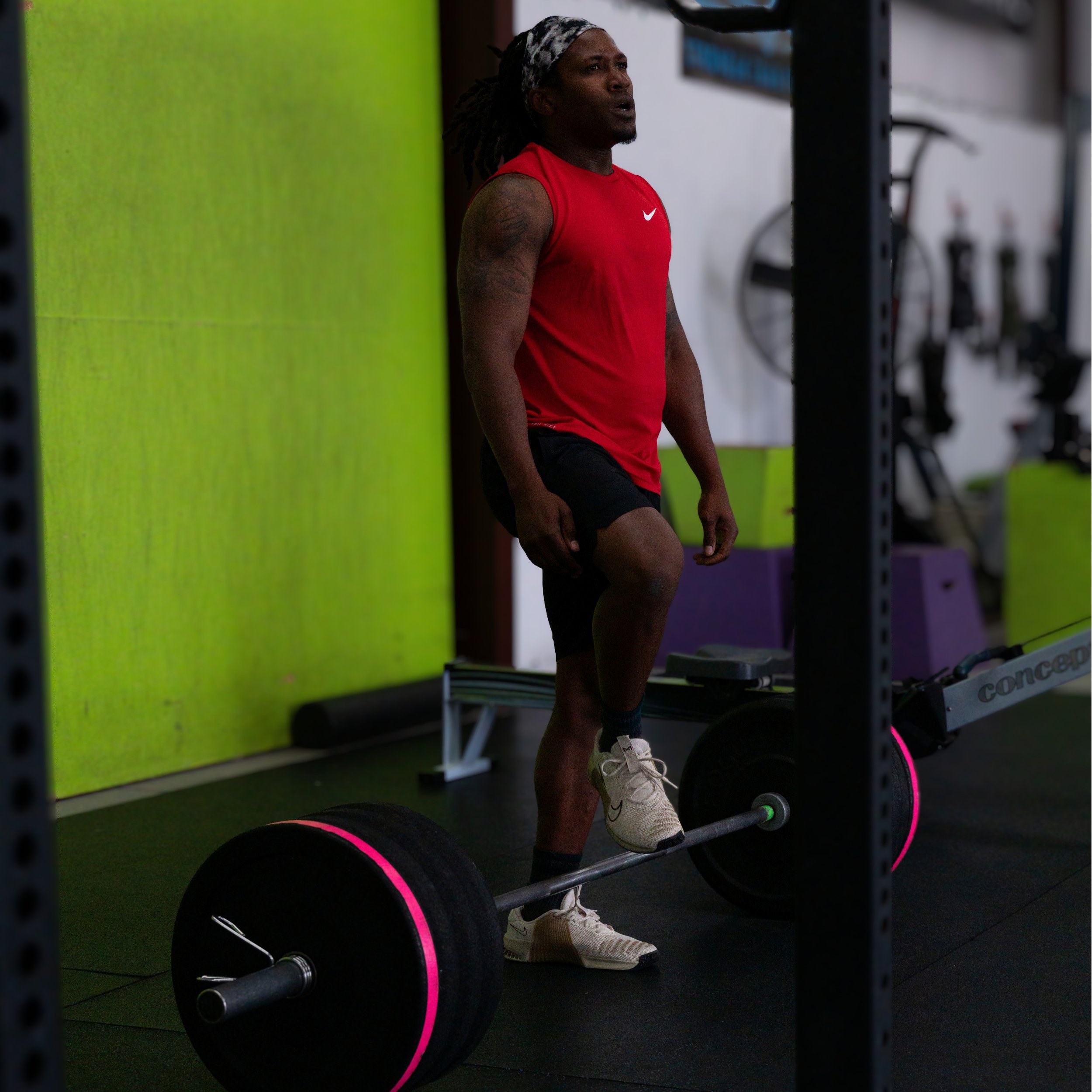 A person in a red sleeveless shirt and black shorts with white sneakers is leaning against a green wall in a gym. They are surrounded by black weights and a black vertical support structure. The person has dreadlocks and tattoos on their right arm, and their expression appears focused.