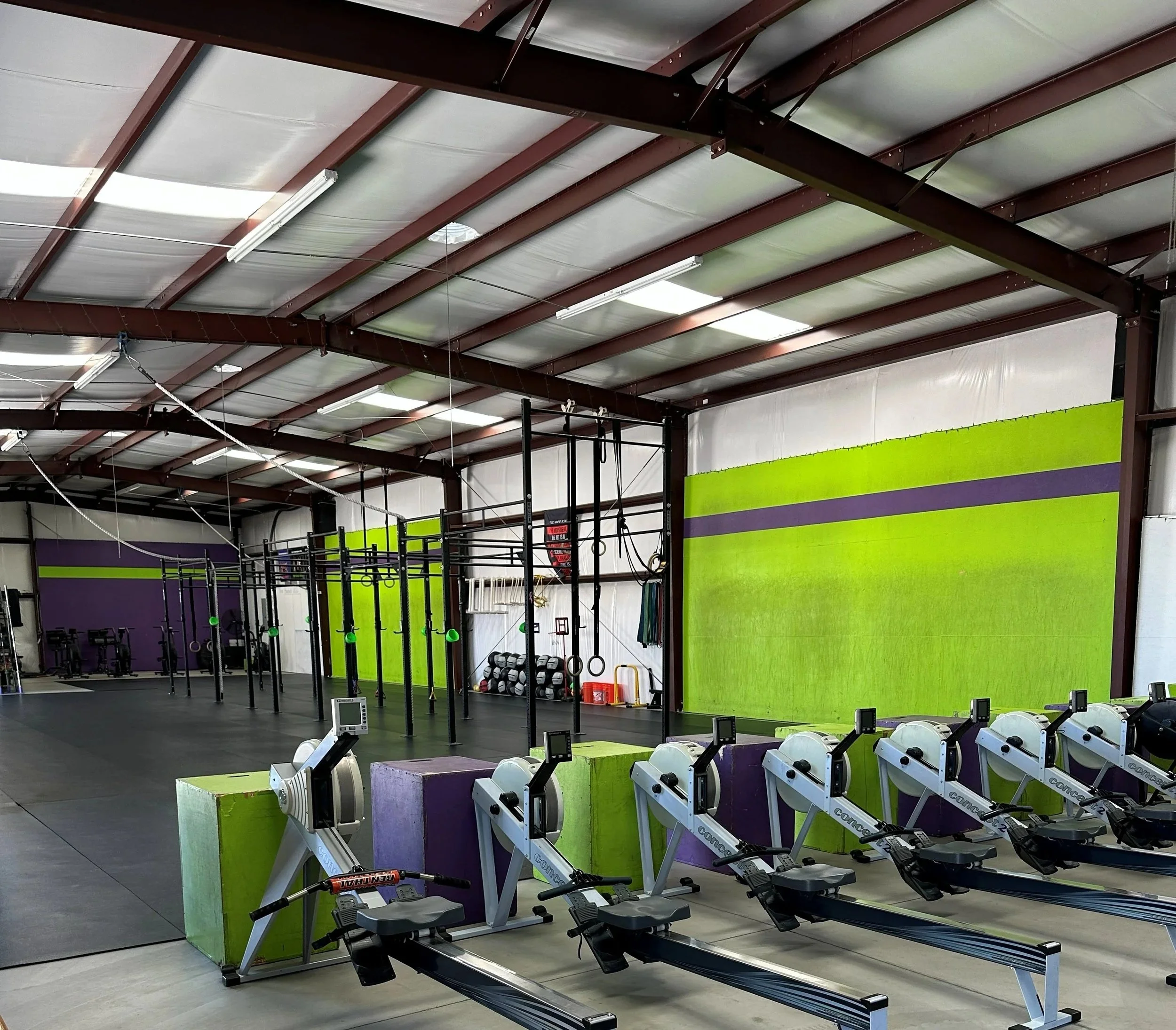 An indoor gym or fitness center with rowing machines in the foreground and a large green and purple wall in the background. The ceiling is made of metal beams and insulated panels, with fluorescent lights.