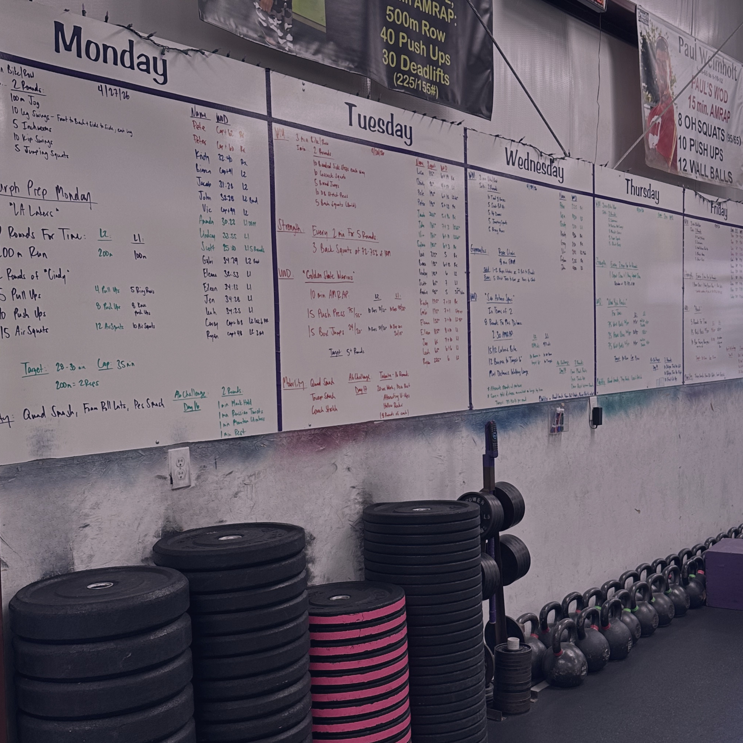 A gym wall decorated with workout schedule and motivational posters, with stacks of weight plates and kettlebells on the floor, and weights organized on racks.