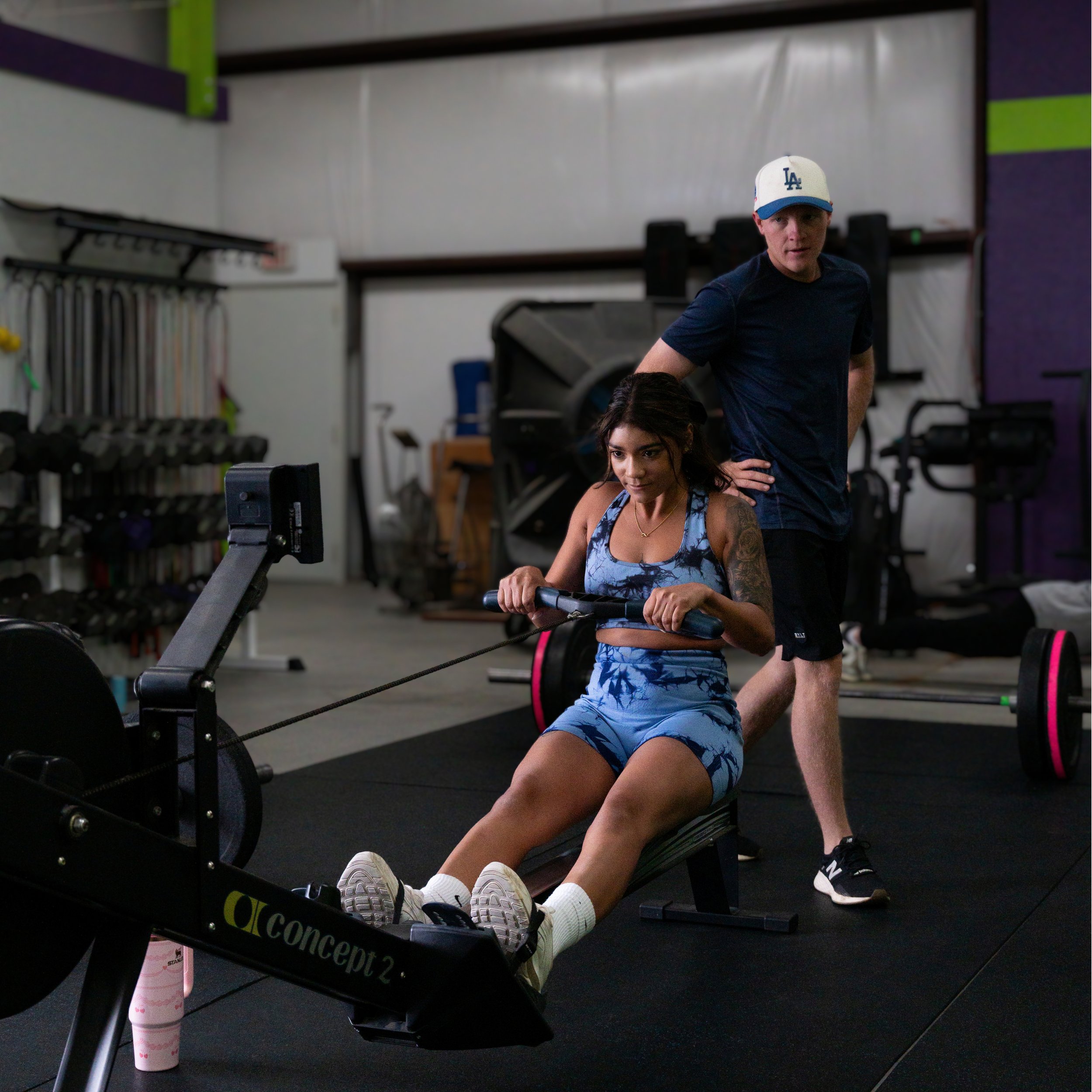A young woman in workout clothes sitting on a rowing machine in a gym, with her feet on the footrests and hands gripping the handle. A man standing behind her appears to be coaching or giving instructions. The gym has various fitness equipment in the background, including weights and exercise machines.