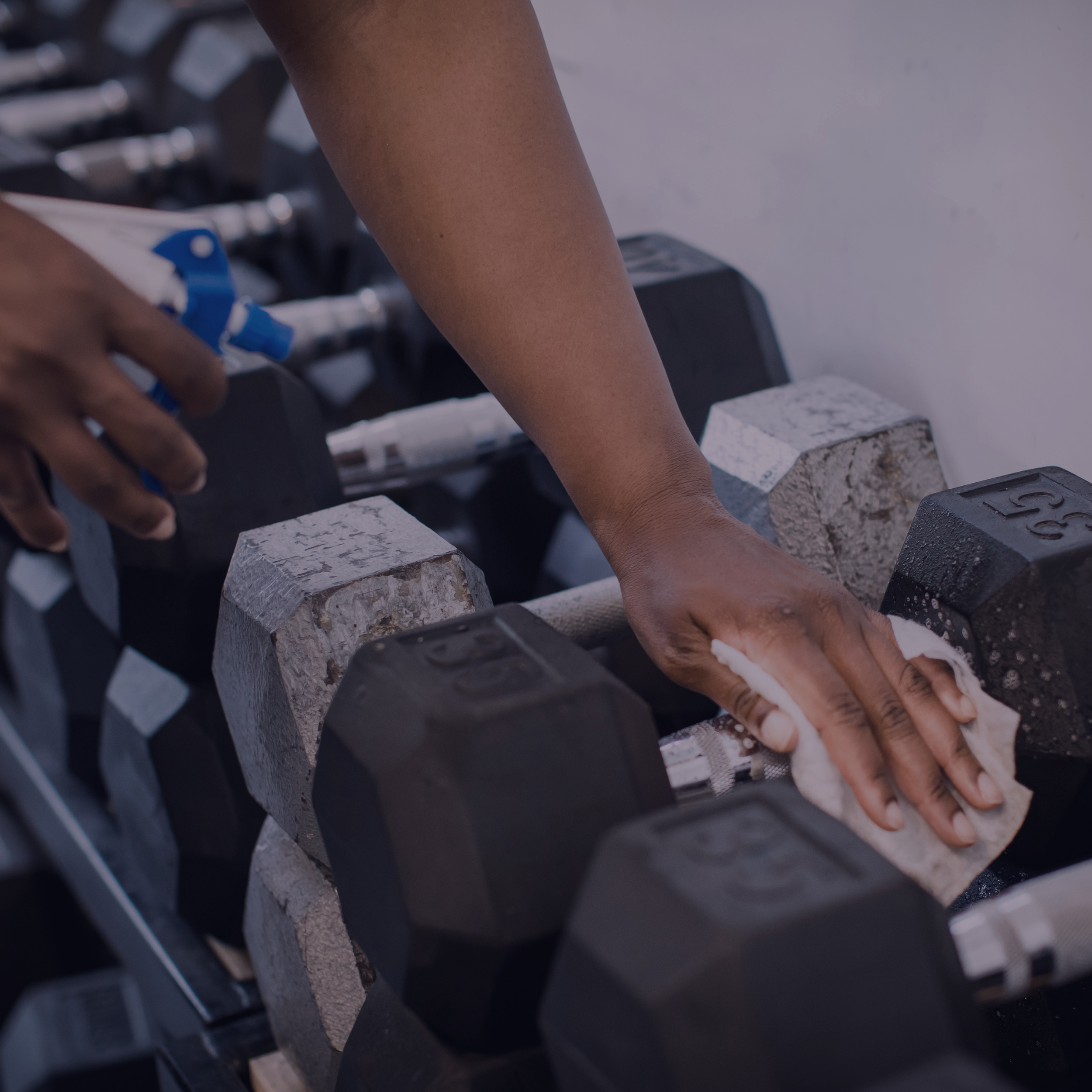 A close-up of a person's hand resting on a set of black and gray rubber weights, with a hand towel underneath their palm, in a gym environment.