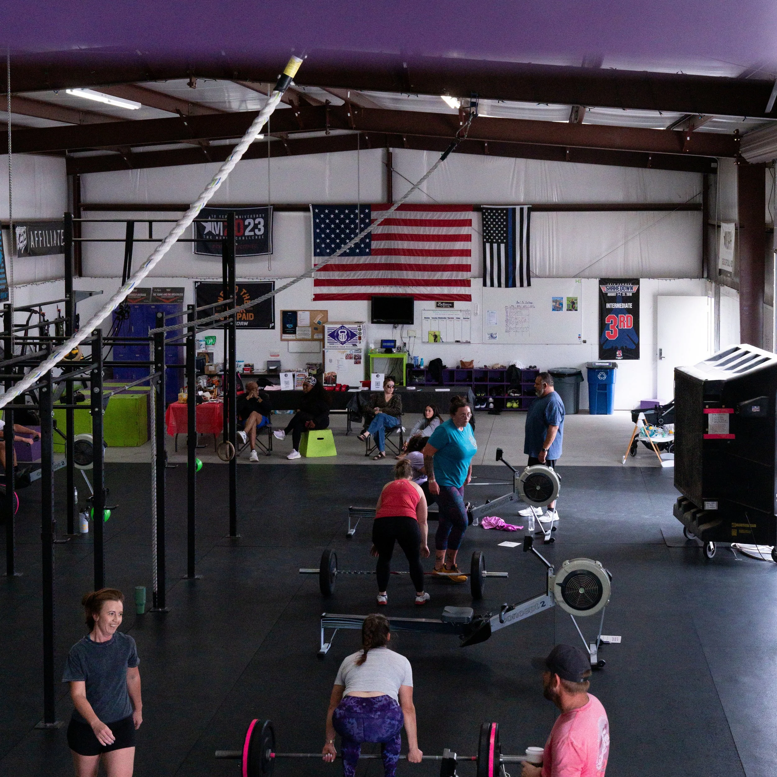 People working out in a gym, lifting weights and using exercise equipment, with American flags hanging on the wall.