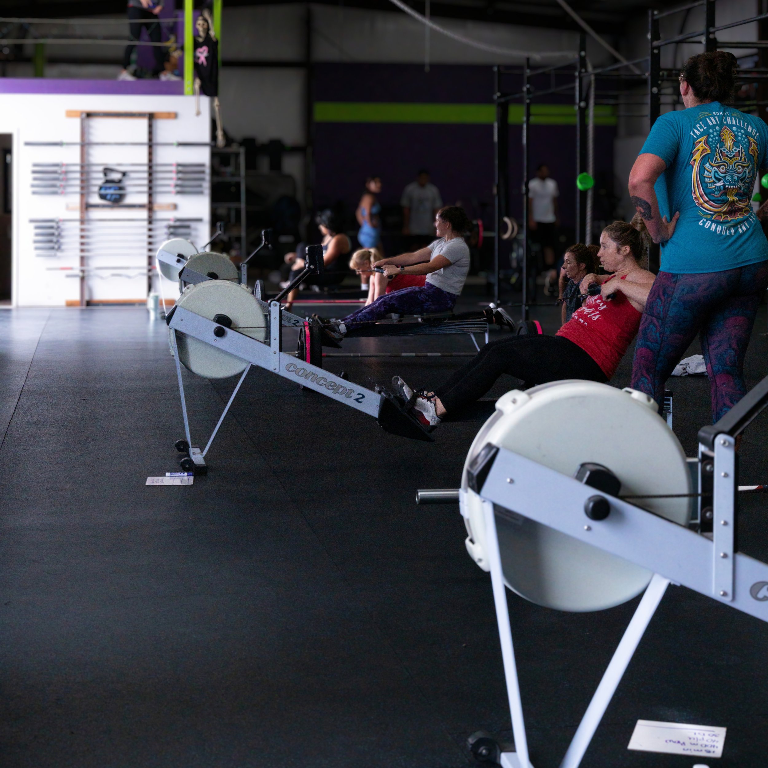 Gym interior with people exercising on rowing machines, a person on the right wearing a blue T-shirt and several others seated or standing, workout equipment visible, dark walls with green accents.