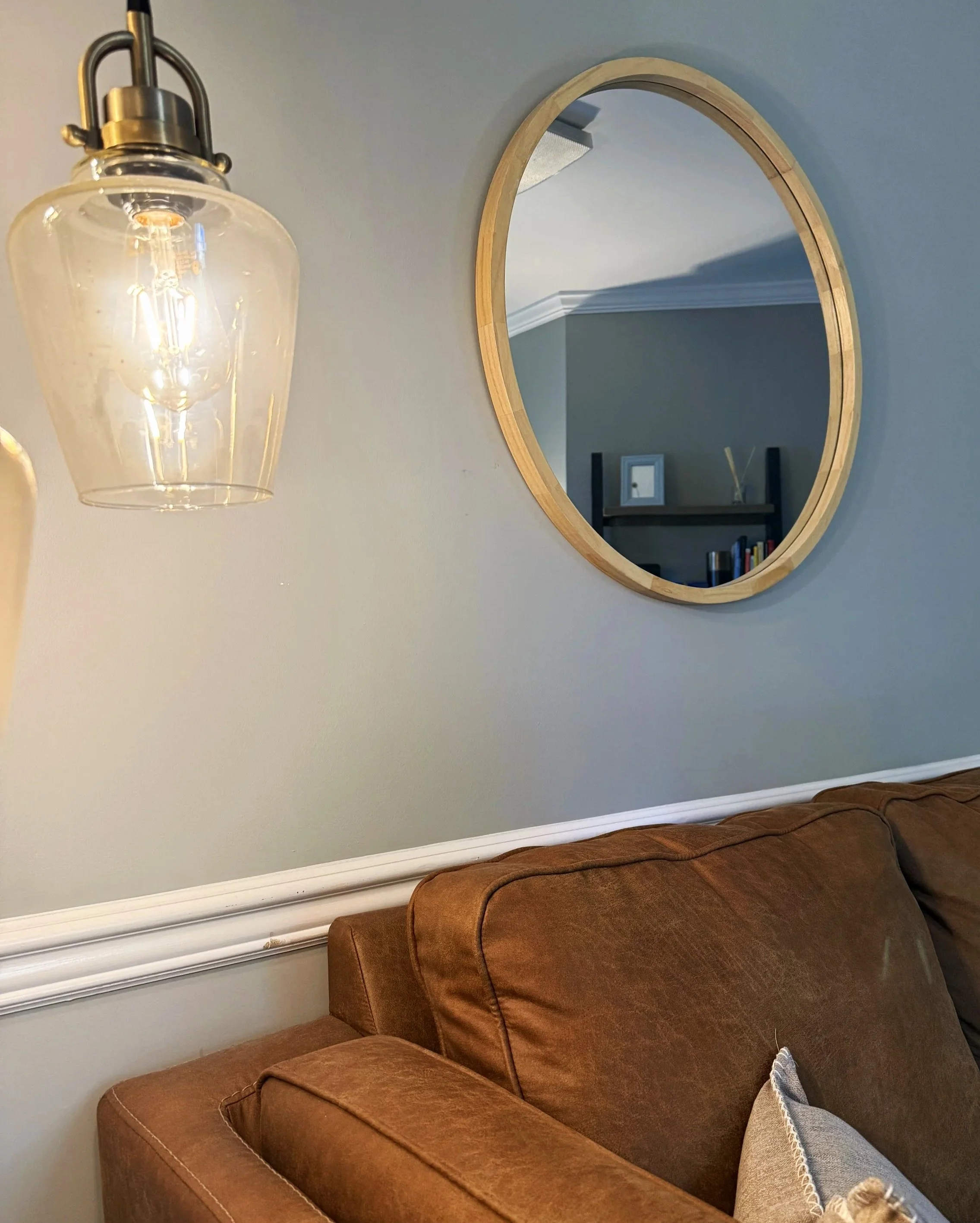 A room with a brown leather sofa, a round wooden-framed mirror on a gray wall, a glass light fixture, and a white chair rail.