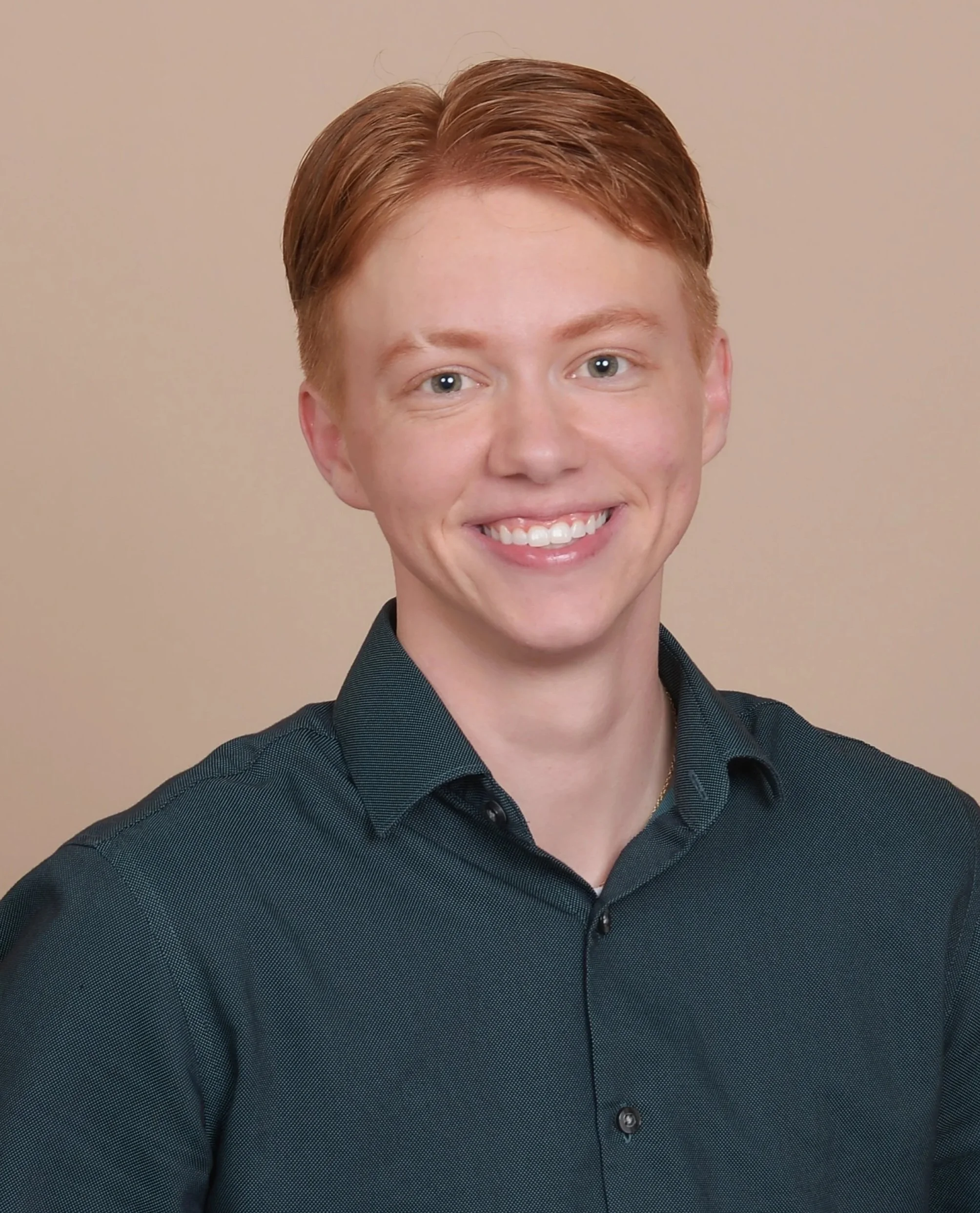 Close-up of a young person with short red hair, smiling, wearing a dark teal collared shirt, against a plain beige background.