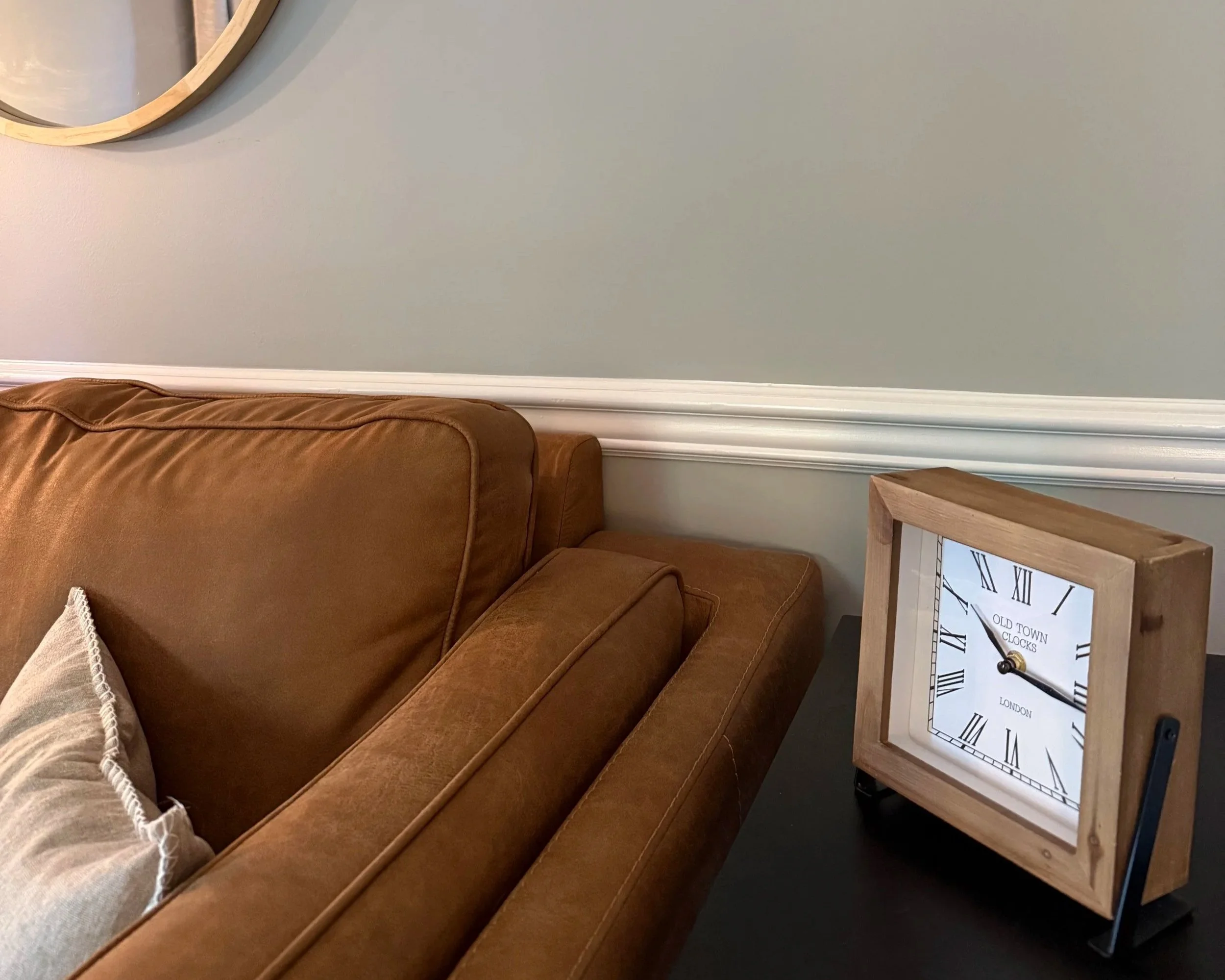Living room corner with a brown leather couch, a beige blanket, a wooden side table, and a large wall clock displaying Roman numerals.