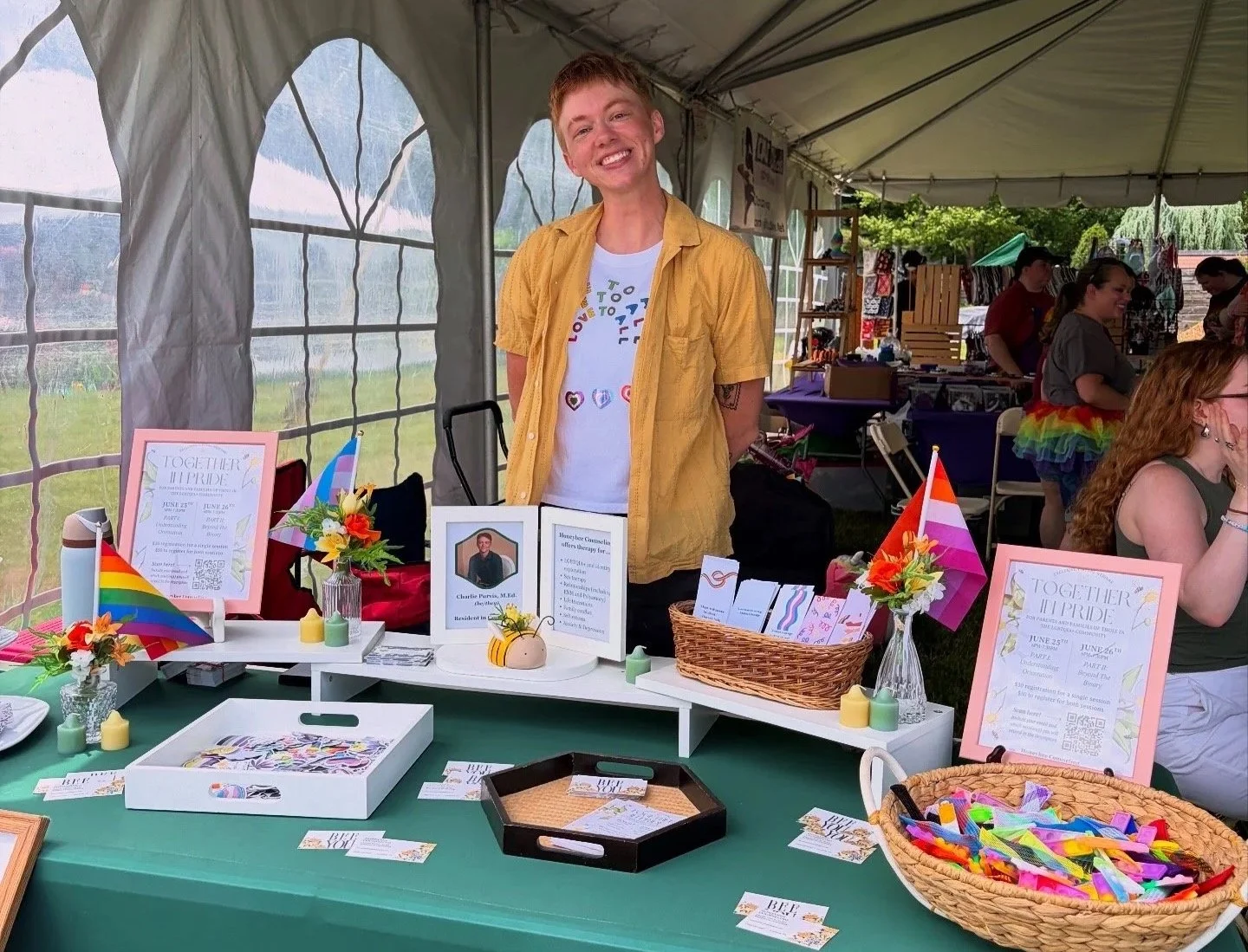 A smiling person wearing a yellow shirt standing behind a table decorated with pride flags, flowers, and informational flyers, at an outdoor pride event under a tent.